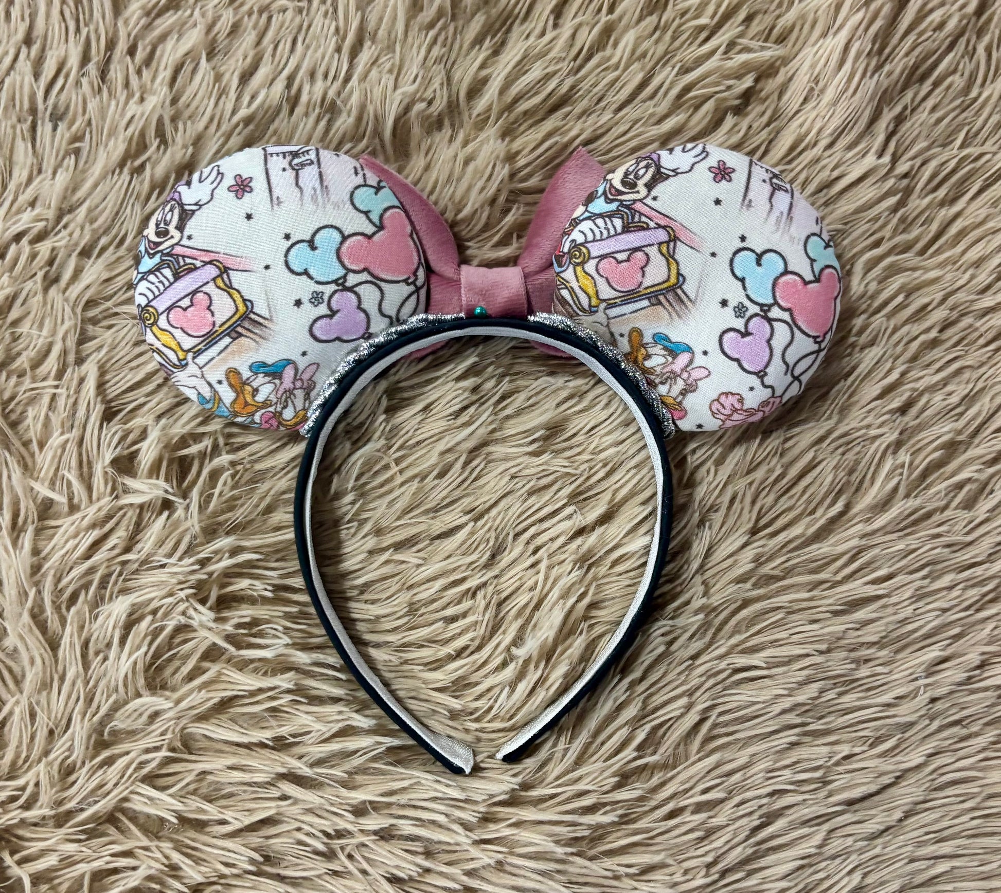 Back view of A pair of handmade ear headbands with a large dusty pink velvet bow, featuring printed park themed patterns on the ears and resting on a beige faux fur surface.