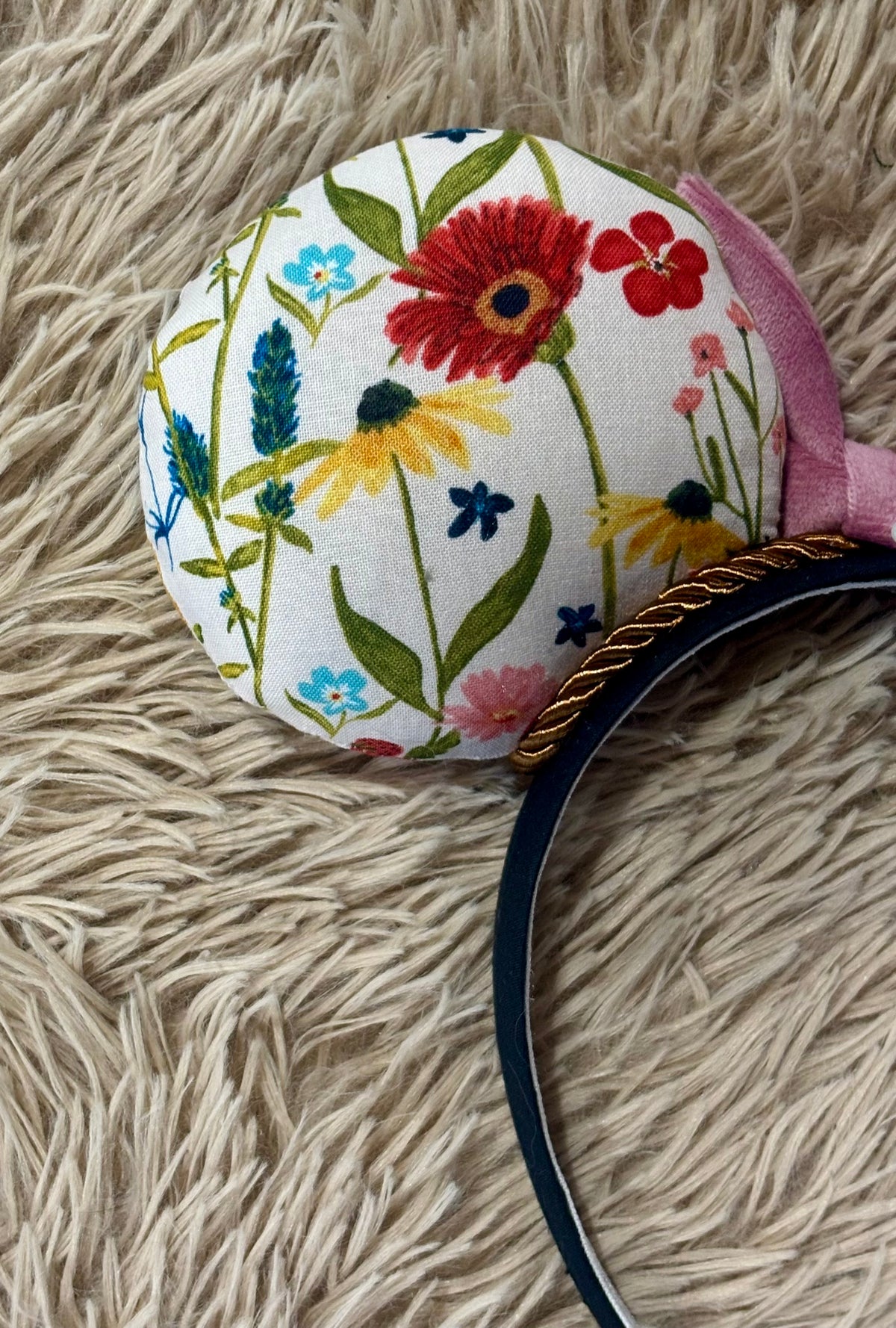 A close-up of a round fabric-covered headband with a colorful wildflower print on a white background, featuring a pink ribbon accent and a black band with a gold rope detail, resting on a beige shag rug.