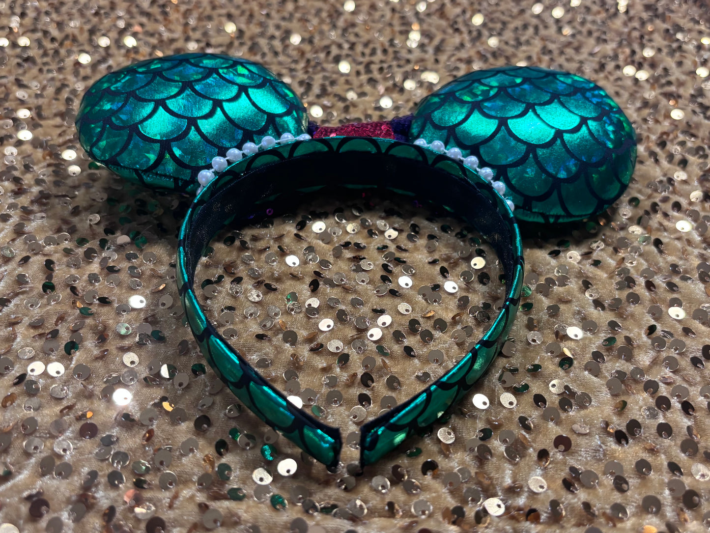 Mermaid-themed headband with teal scales and a purple sequin bow with a red middle and a dinglehopper middle piece. Pearl garland trim along inner ears m on a matching mermaid scales headband. Back and inner headband view.