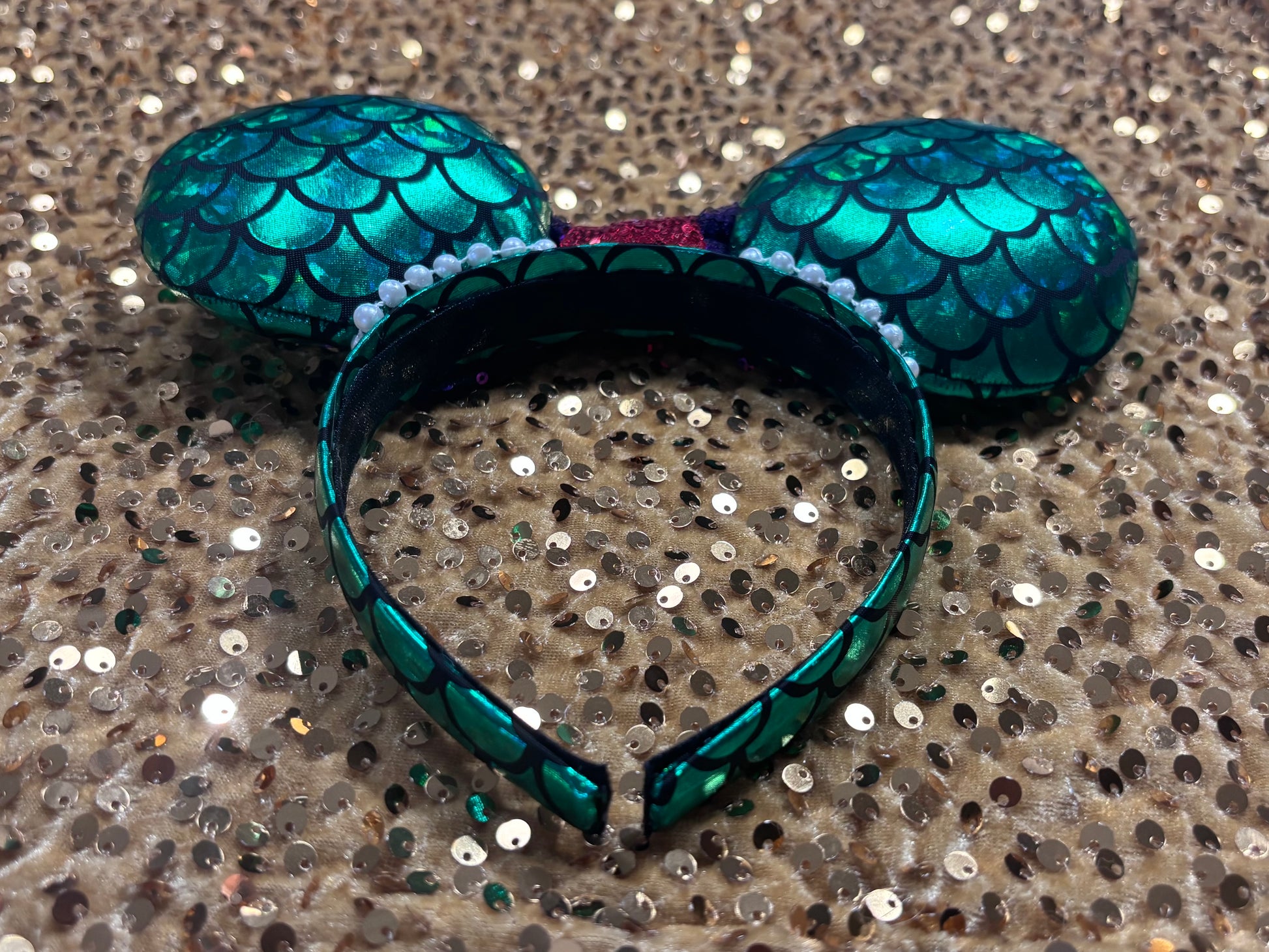 Mermaid-themed headband with teal scales and a purple sequin bow with a red middle and a dinglehopper middle piece. Pearl garland trim along inner ears m on a matching mermaid scales headband. Back and inner headband view.