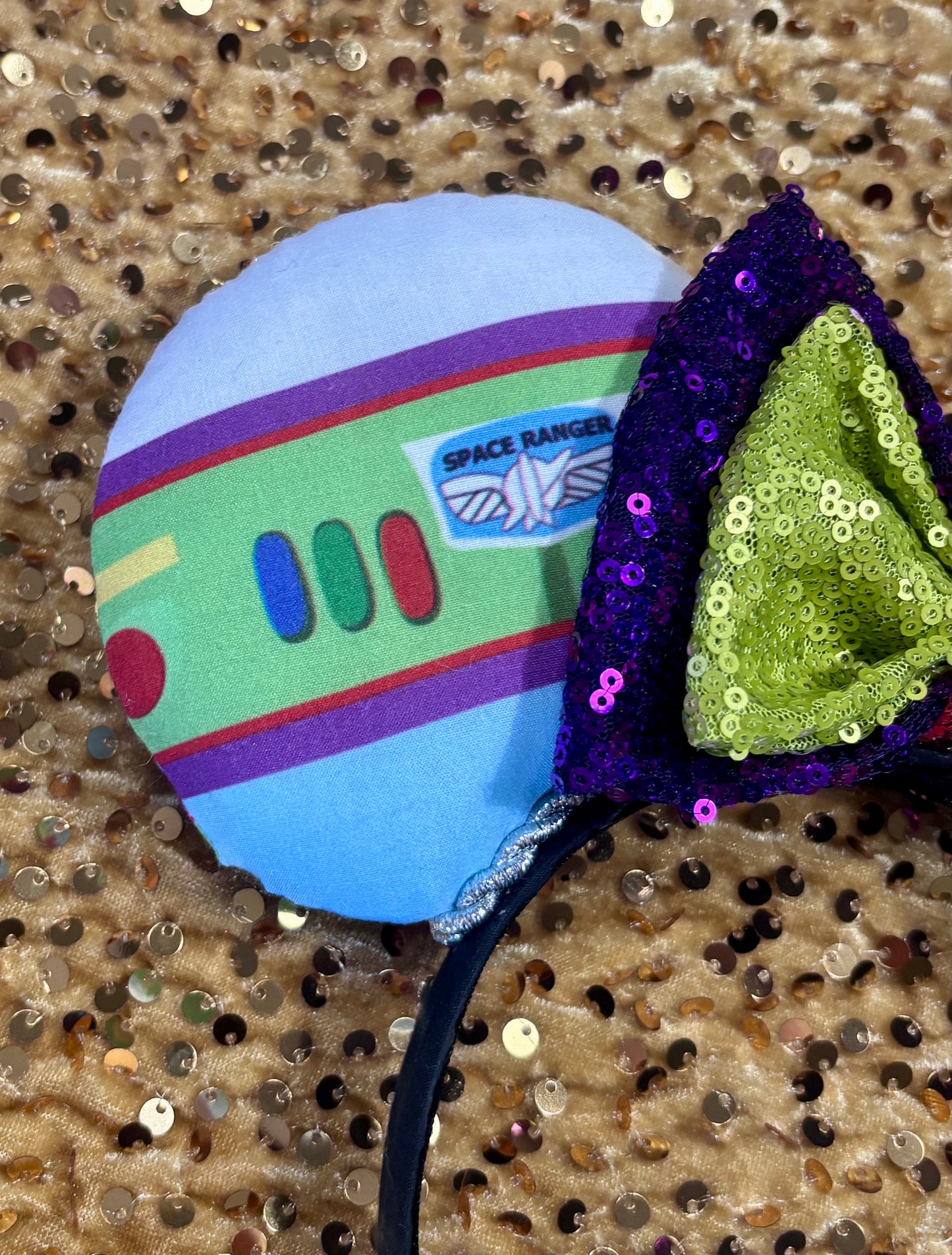 Close up of front left mouse ear. Space Ranger Mouse Ear headband with new improved bow. Handmade and ready to ship with Space ranger fabric on both the front and the back of mouse ears. The set has a silver twisted trim along the inner ears and a new sequin bow that is purple, lime green and red in sequin. Ears are on a black poly cotton universal sized headband.