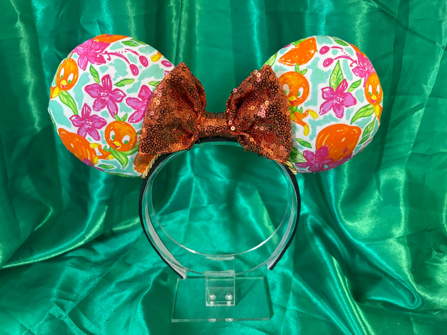 A pair of handmade mouse ears with an orange and pink floral pattern on a black headband, featuring sequin trim and a bow.
