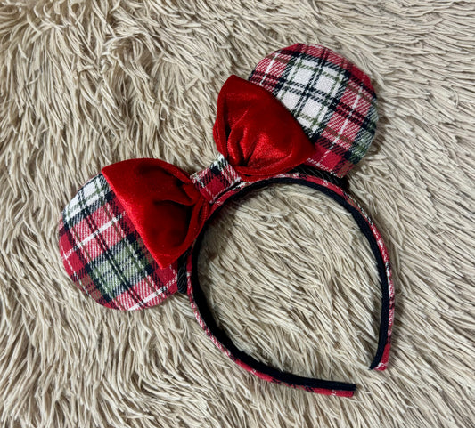 A plaid ear headband with red velvet bows, displayed on a textured beige faux fur background.