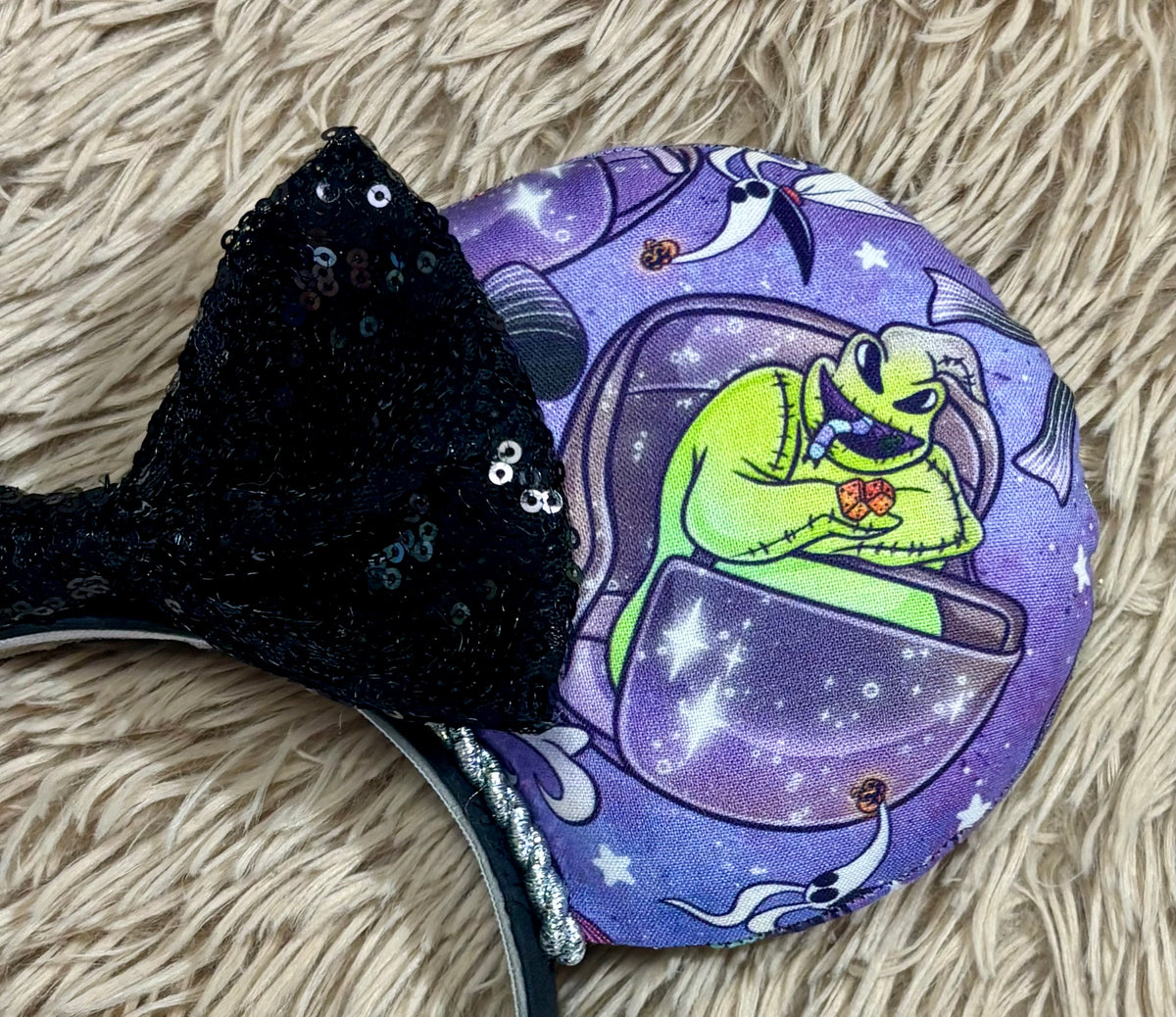An ear headband accessory with a black sequined bow, featuring a purple HM and NBC themed print with a cartoon character in a buggy.