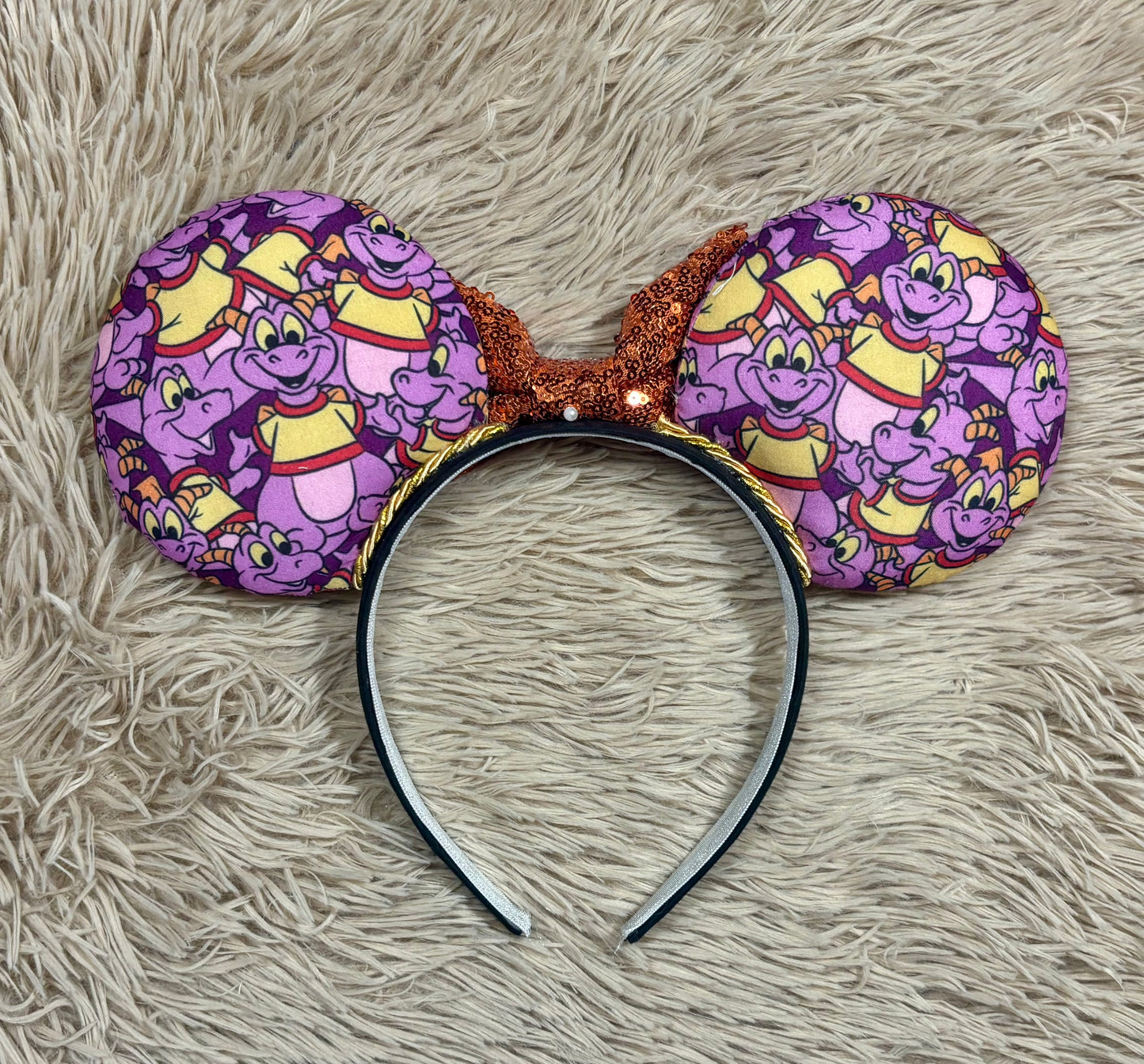 A pair of dragon style ears with a black headband, featuring a purple and yellow cartoon pattern on the ears and an orange sequined bow in the center. Back view of ears.
