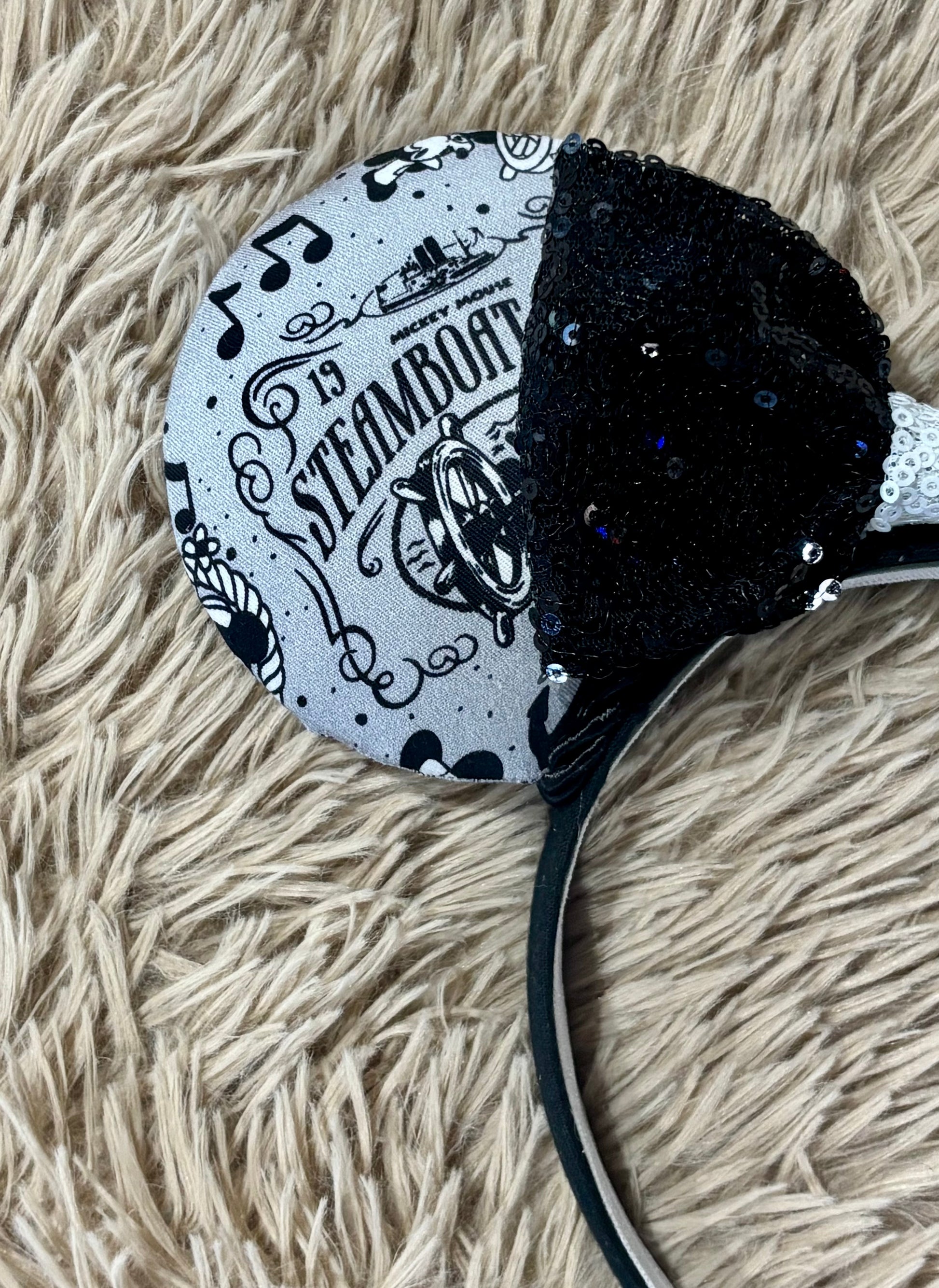 Close up of Mouse-themed ear headbands featuring a black and white patterned fabric with sequins, a black sequined bow with a white sequin middle, and a white headband, displayed on a beige faux fur background.