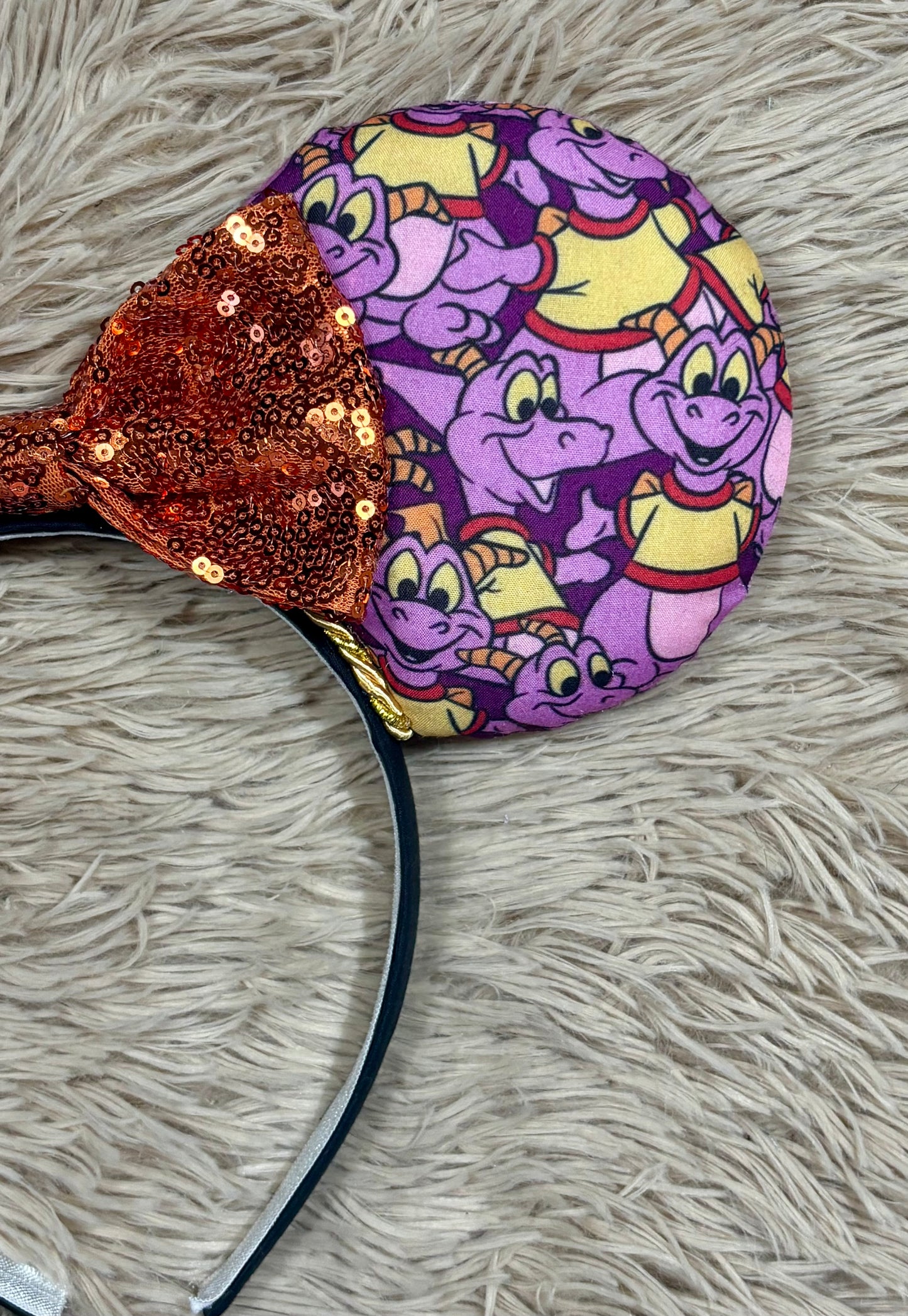 A pair of dragon style ears with a black headband, featuring a purple and yellow cartoon pattern on the ears and an orange sequined bow in the center