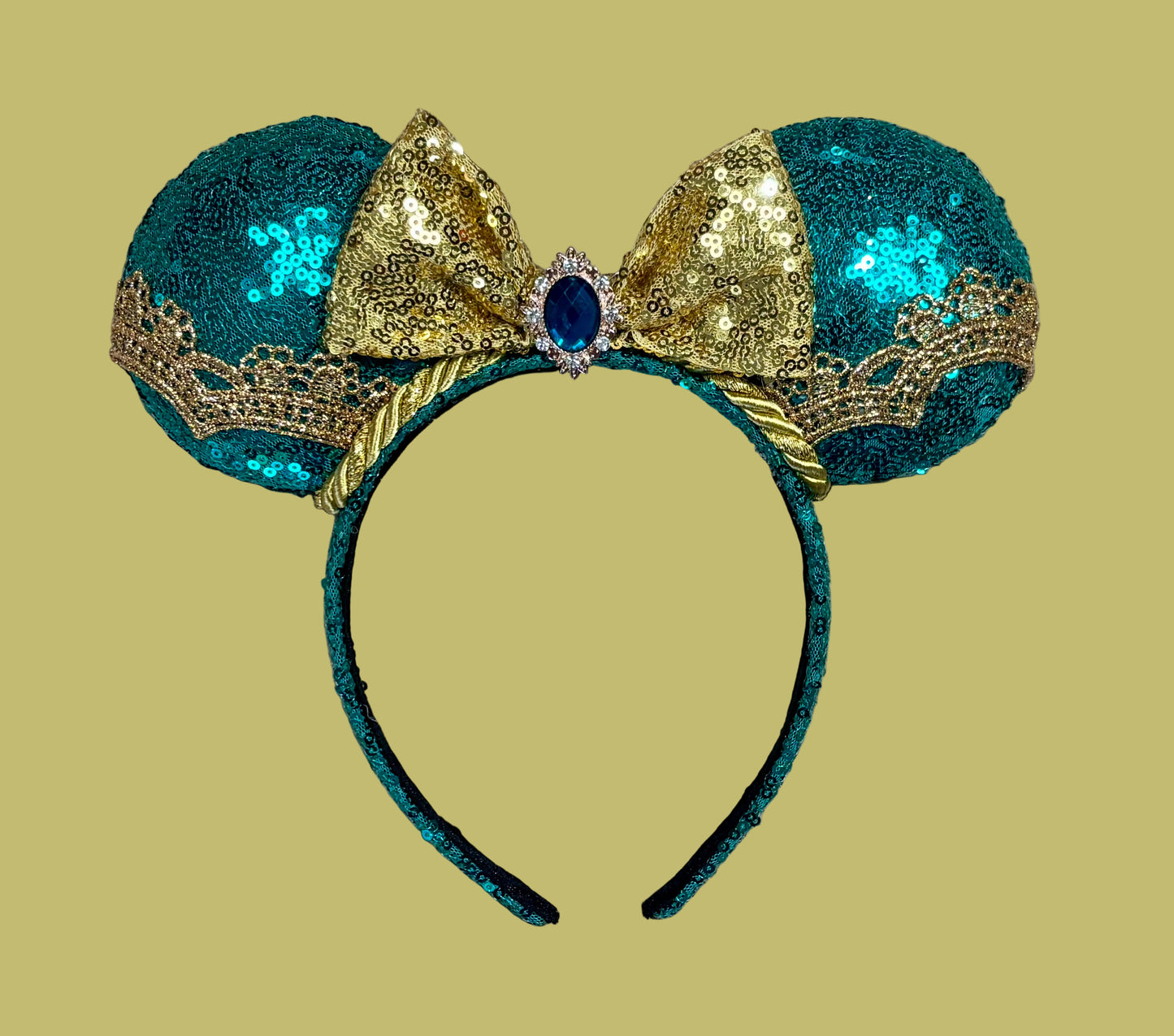 A pair of sequined mouse ears headband featuring teal and gold sequins, a large gold sequin bow with a blue gemstone center, and gold lace trim.