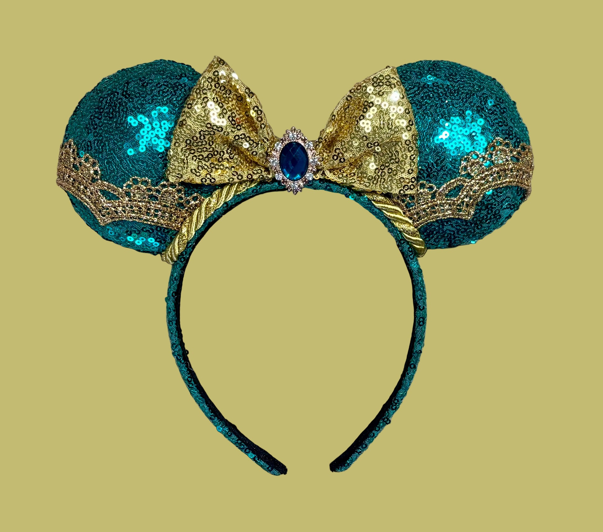 A pair of sequined mouse ears headband featuring teal and gold sequins, a large gold sequin bow with a blue gemstone center, and gold lace trim.