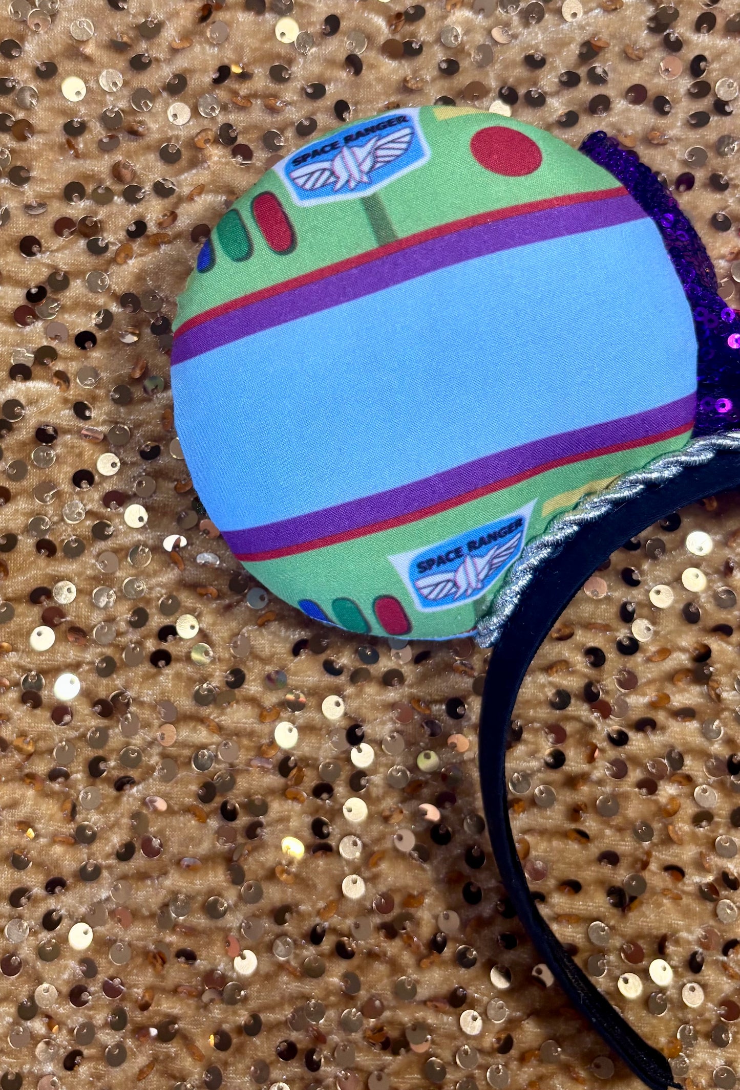 Space Ranger Mouse Ear headband with new improved bow. Handmade and ready to ship with Space ranger fabric on both the front and the back of mouse ears. The set has a silver twisted trim along the inner ears and a new sequin bow that is purple, lime green and red in sequin. Ears are on a black poly cotton universal sized headband. Close up of back left mouse ear.