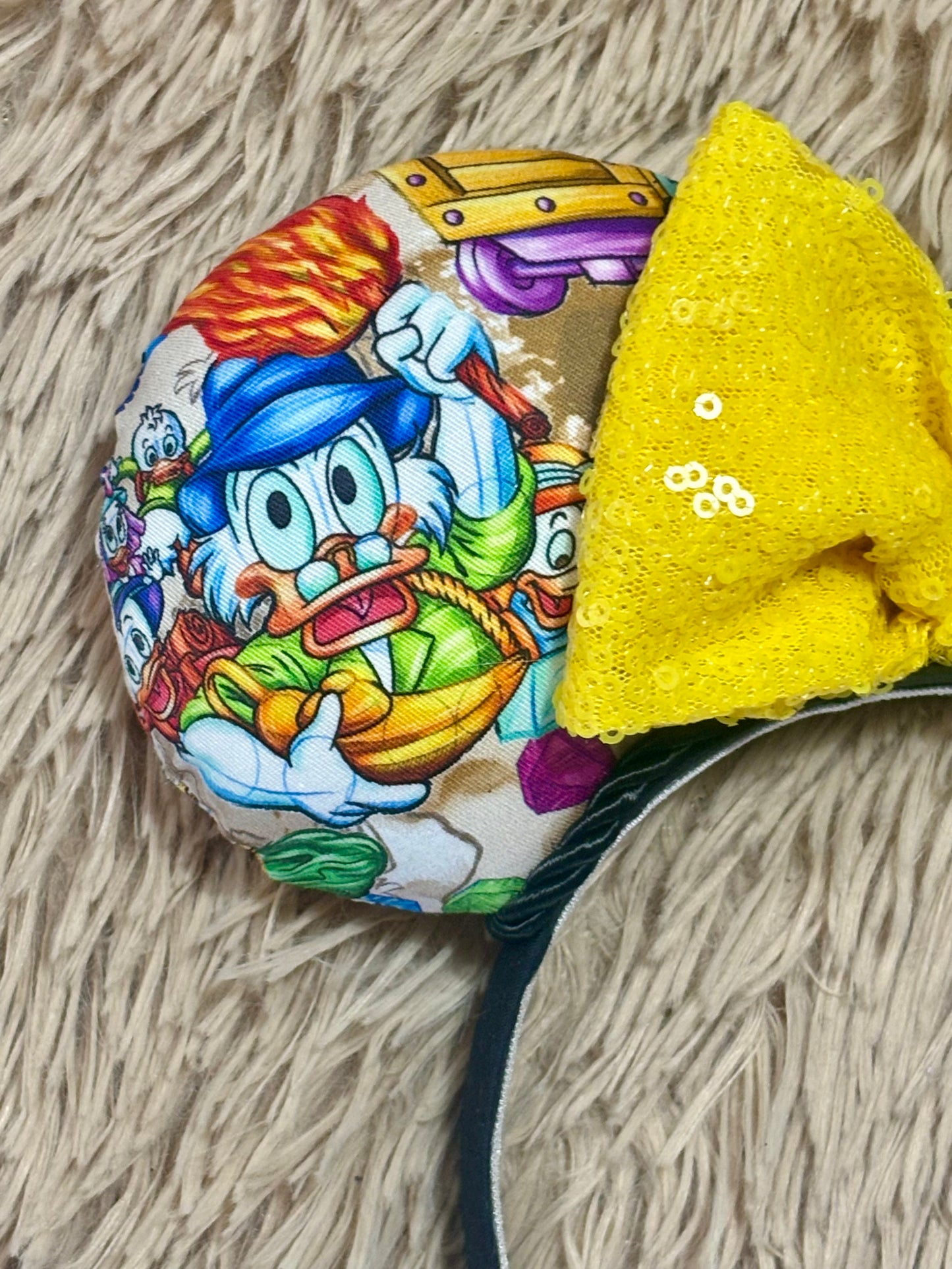 A close-up of the front ear on the headband featuring a colorful cartoon character print and a large yellow sequined bow, resting on a beige shag rug.