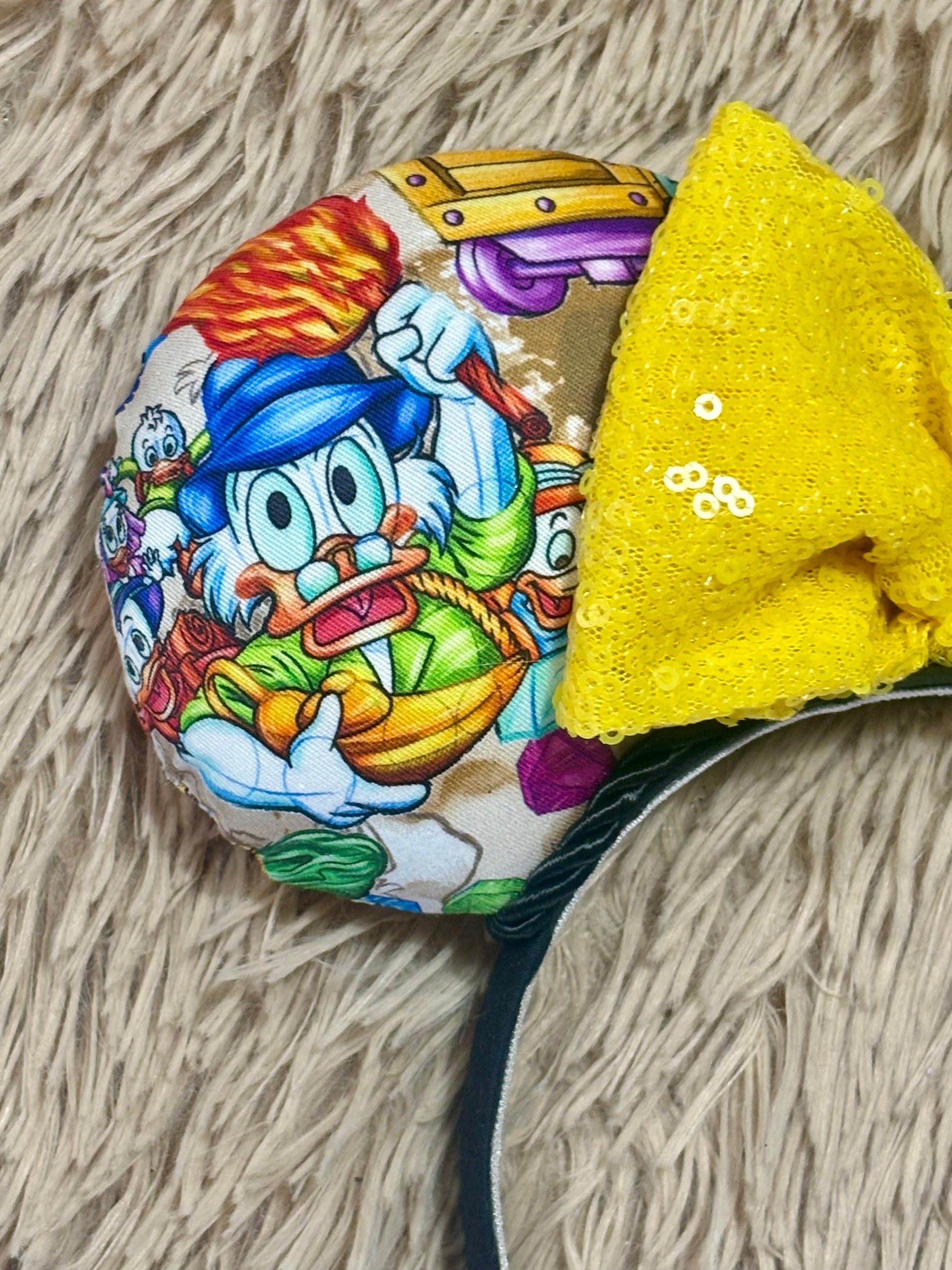 A close-up of the front ear on the headband featuring a colorful cartoon character print and a large yellow sequined bow, resting on a beige shag rug.