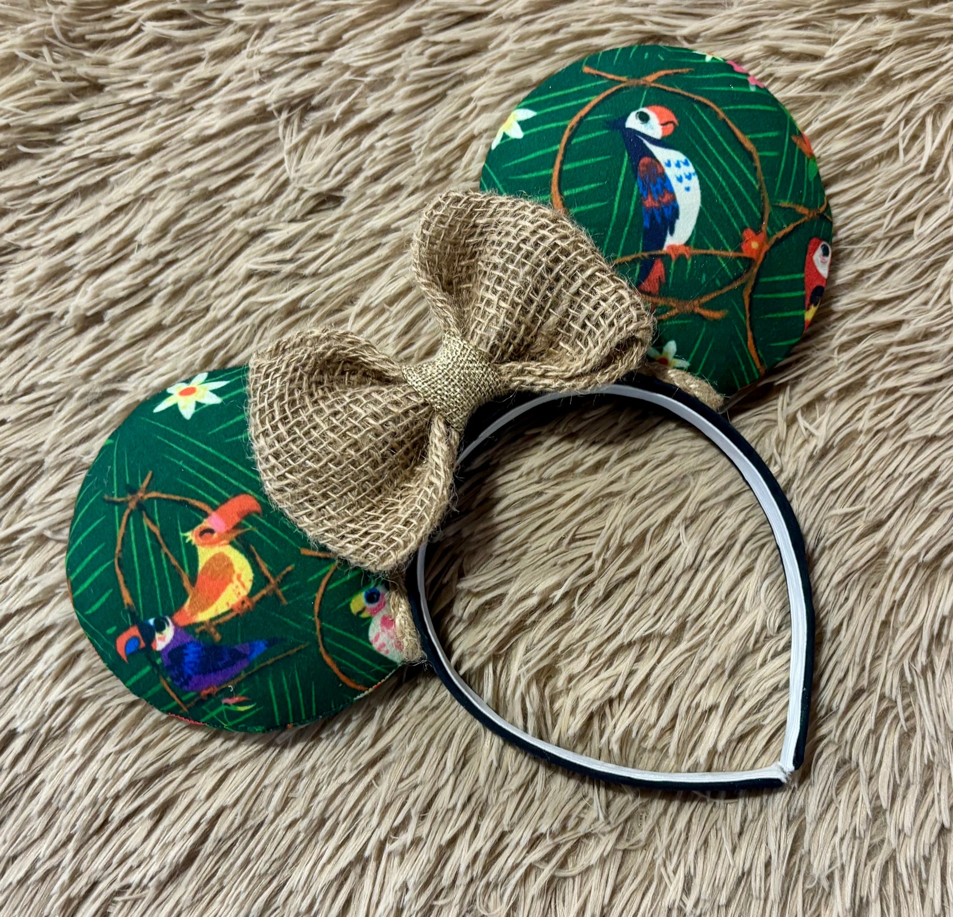 A headband with two round, bird-printed ear-like pieces attached, featuring a large natural burlap bow on the front, photographed on a beige shaggy faux-fur surface.