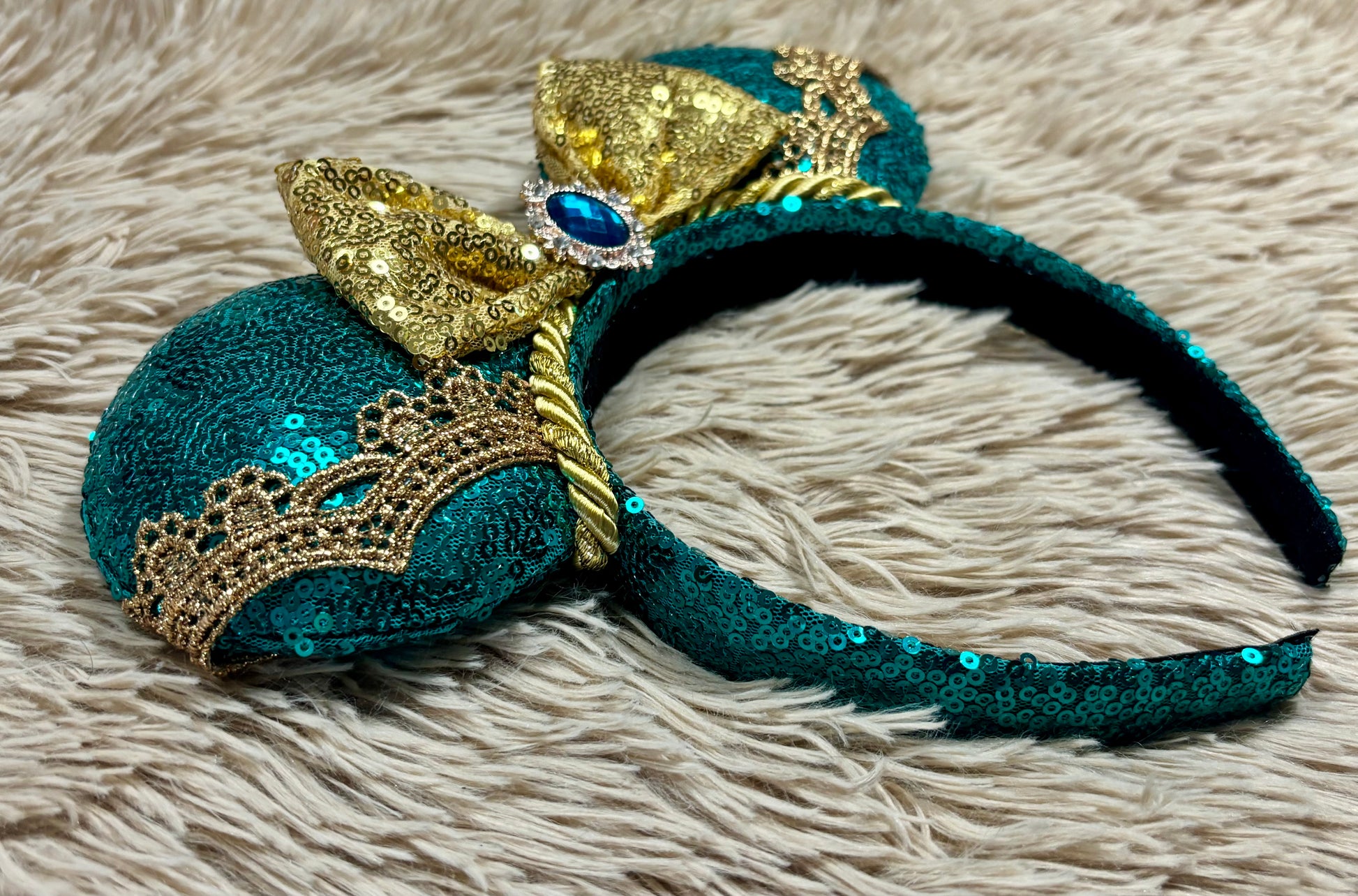 A close-up of a decorative headband with large round ears covered in teal sequins, embellished with gold sequin bows, a gold tiara motif, and a central blue gem, resting on a beige shaggy rug.