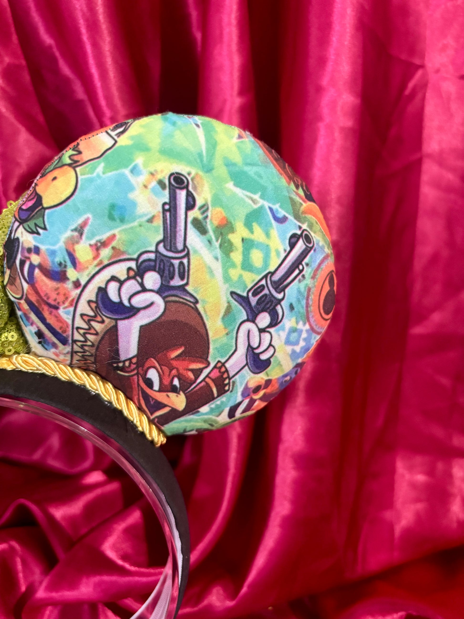 Colorful Handmade mouse ears with a green bow on a red satin background with duck cartoon images on the ears. Close up of back right mouse ear.
