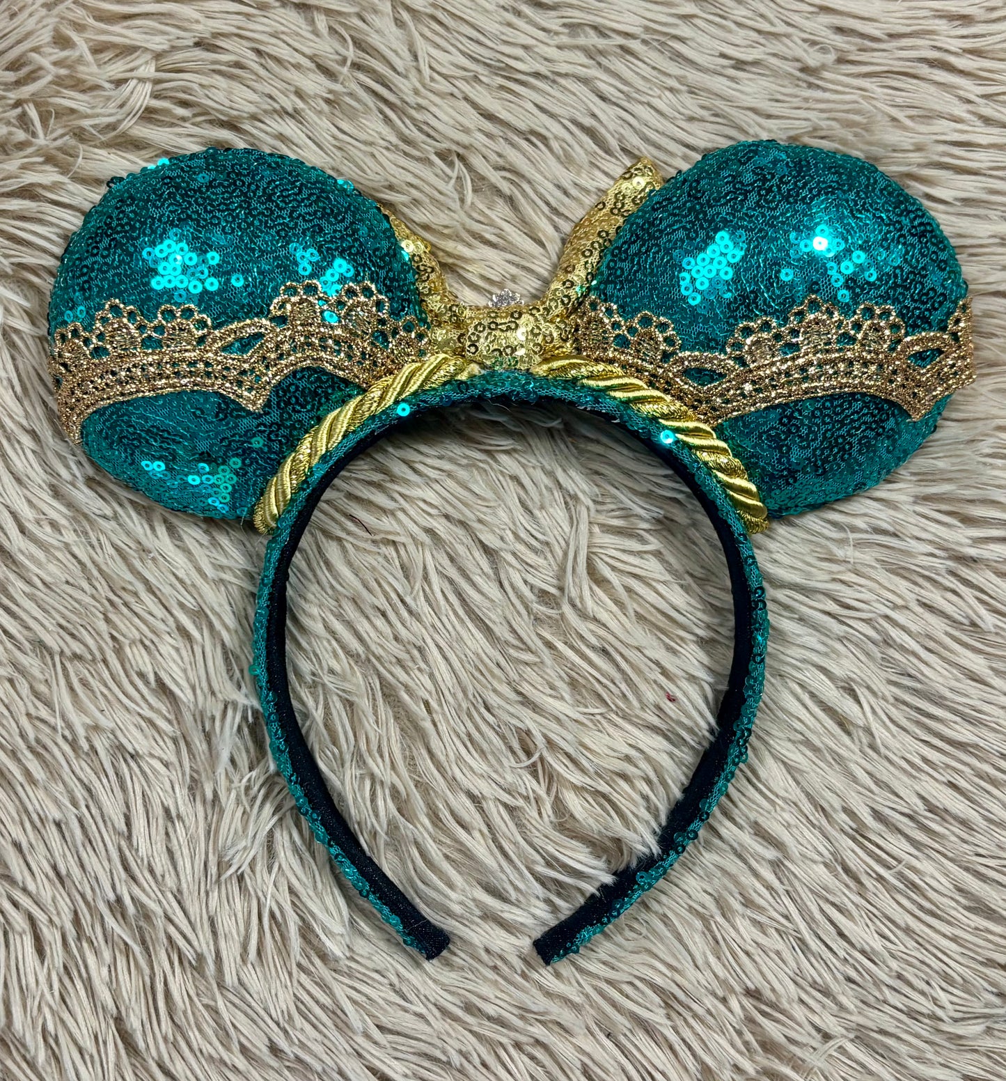 back view of a pair of sequined mouse ears headband featuring teal and gold sequins, a large gold sequin bow with a blue gemstone center, and gold lace trim.