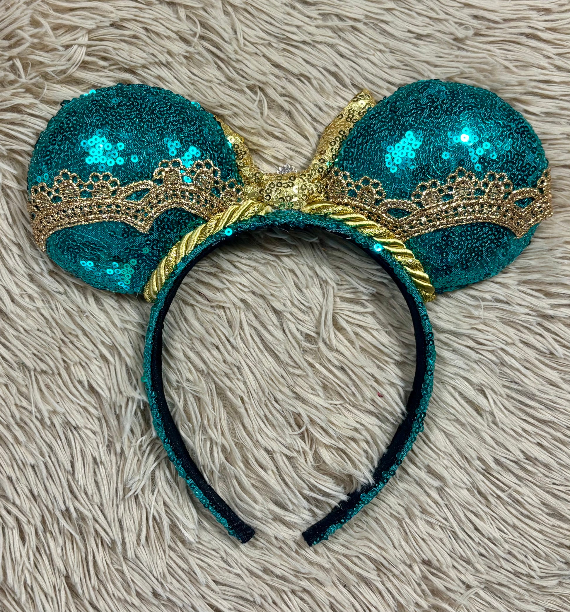 back view of a pair of sequined mouse ears headband featuring teal and gold sequins, a large gold sequin bow with a blue gemstone center, and gold lace trim.
