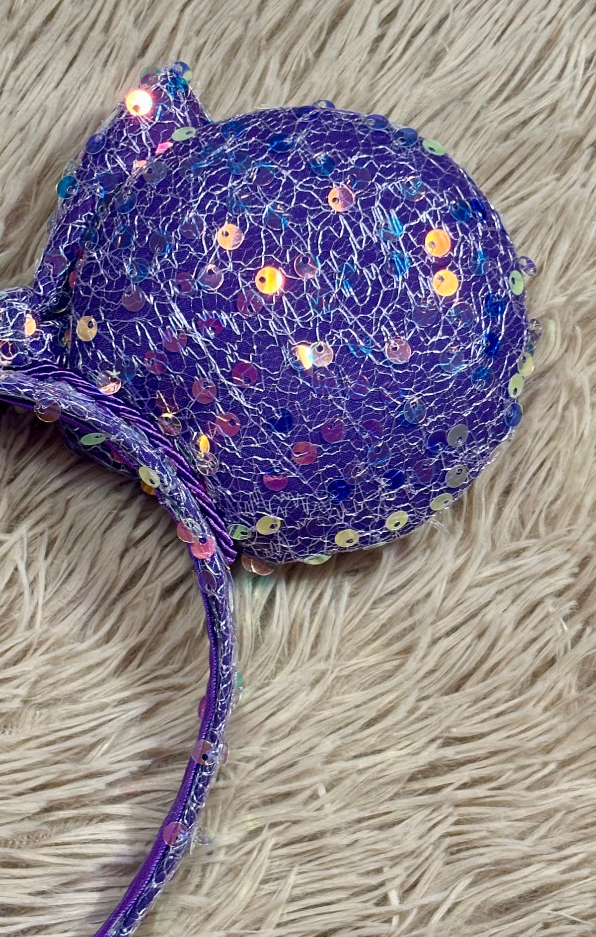 A close-up of a purple sequined headband with an elastic band, resting on a beige shaggy rug.