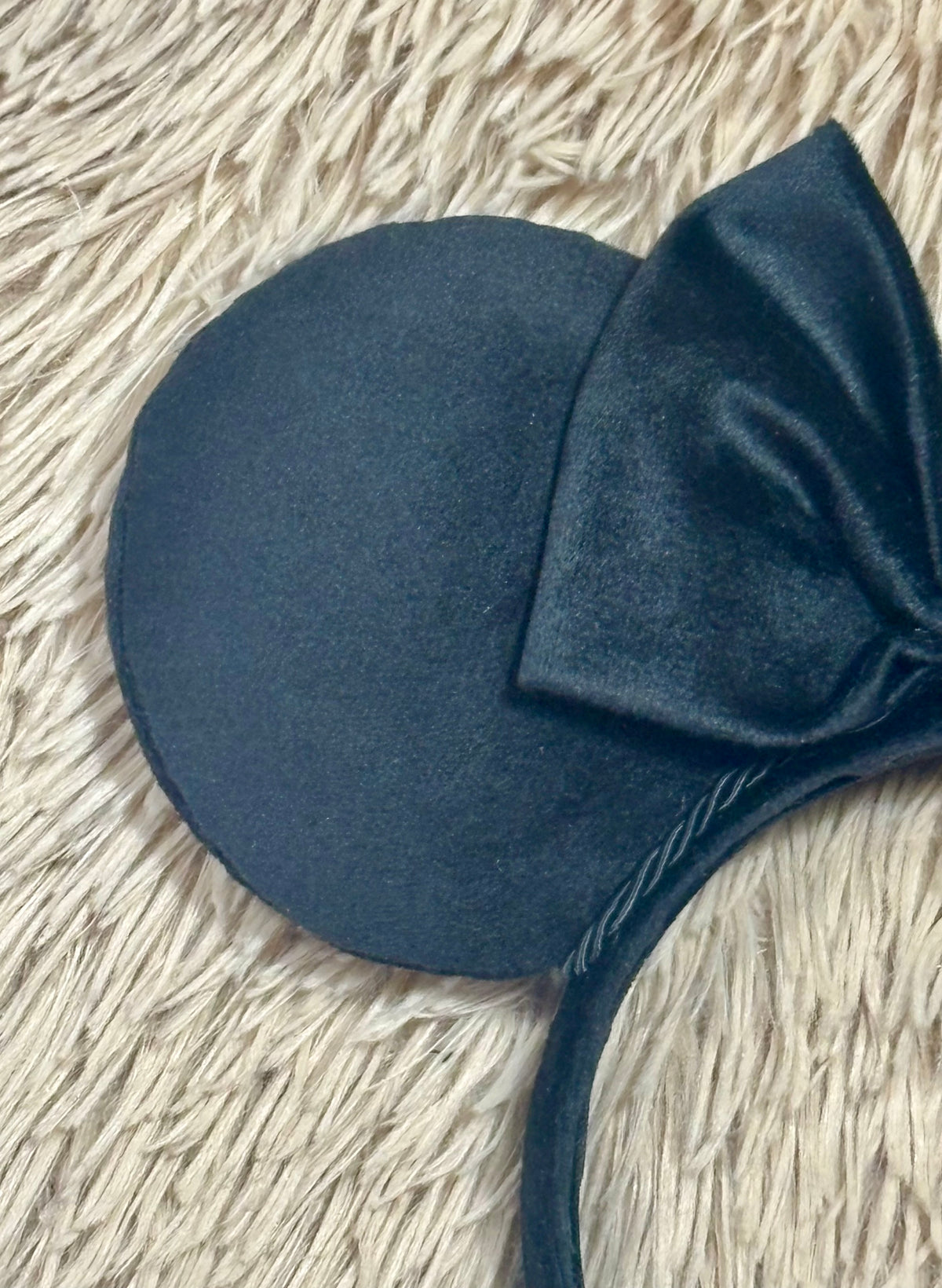 A close-up of a black mouse-ear headband with a large velvet bow, laid on a beige shaggy textured surface.