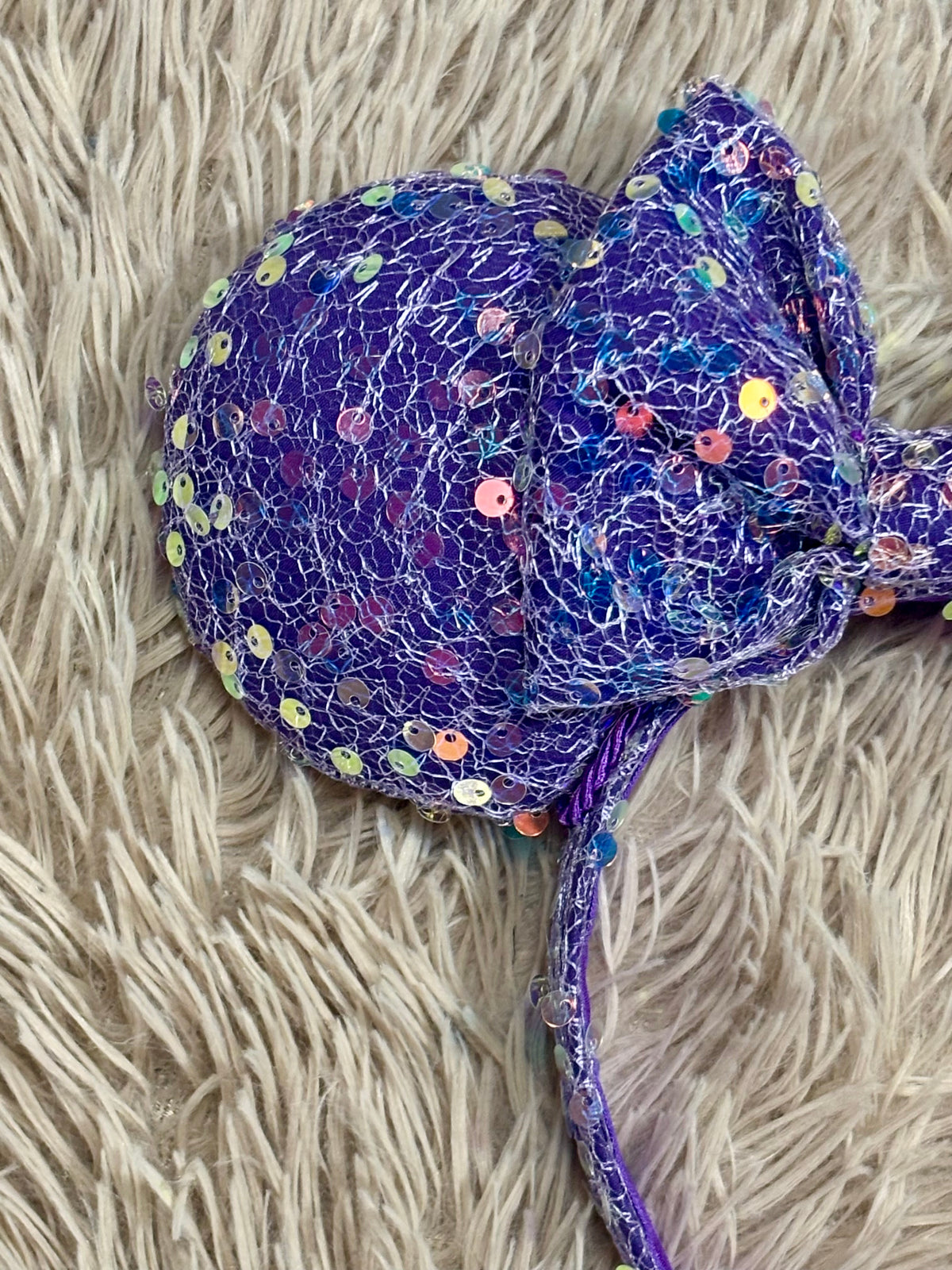 A purple sequined headband with a large bow, laid flat on a beige shag rug.