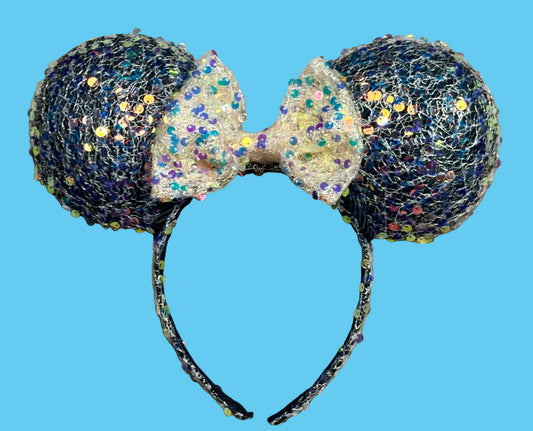 A pair of sparkly ears headband featuring black glitter-covered round ears and a large cream bow with iridescent glitter, displayed on a blue background.