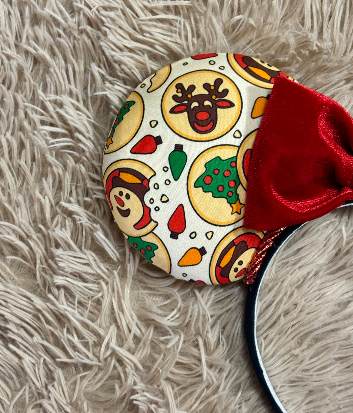 A close-up of a round holiday-themed ear with a white background, colorful cartoon snowman and Christmas tree motifs, and a red plush bow, placed on a shaggy beige rug.