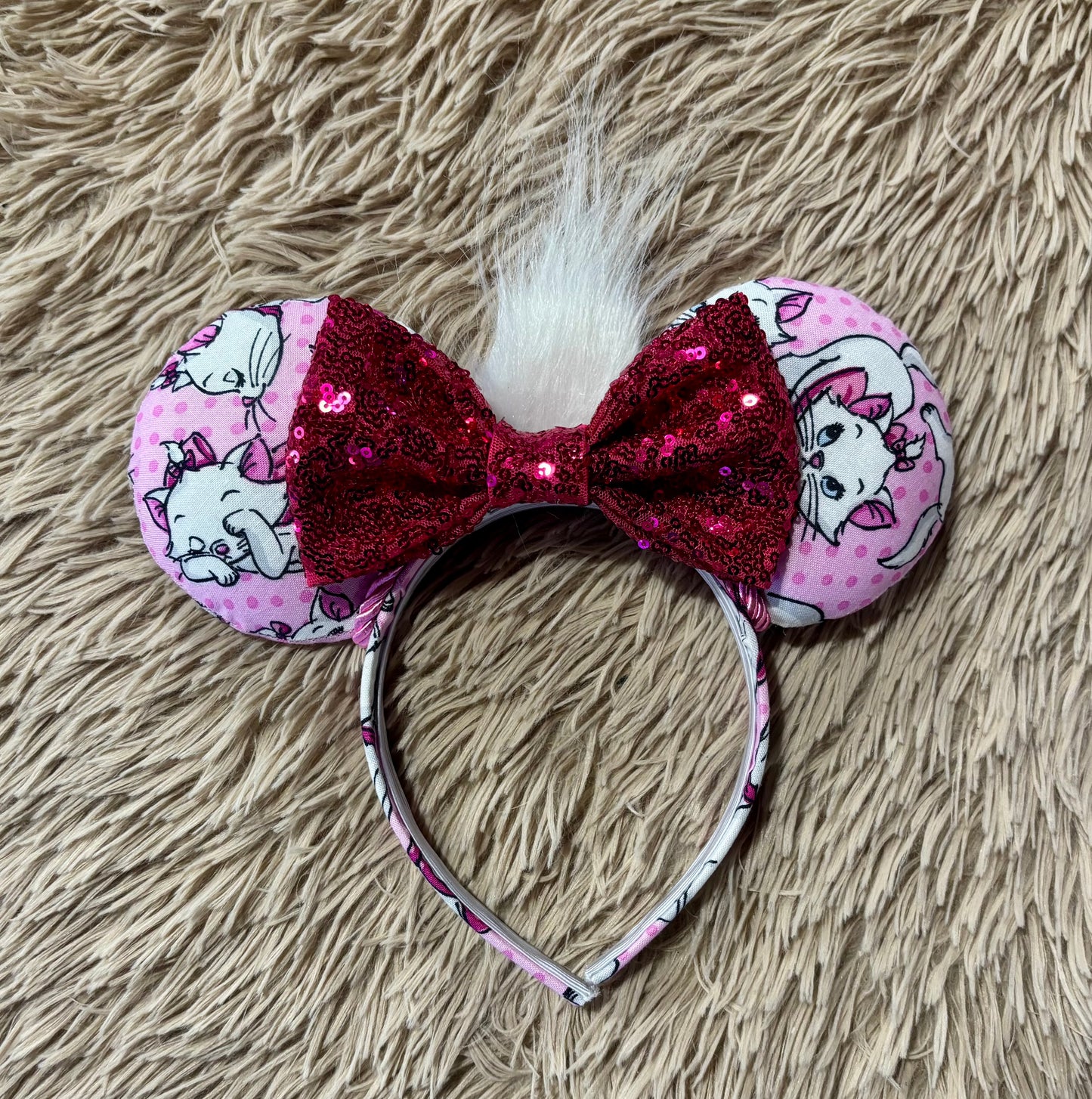 A pair of Marie inspired ears featuring pink fabric with white cartoon cats and pink polka dots, a large hot pink sequined bow, and a white faux fur accent, displayed on a beige shag rug.