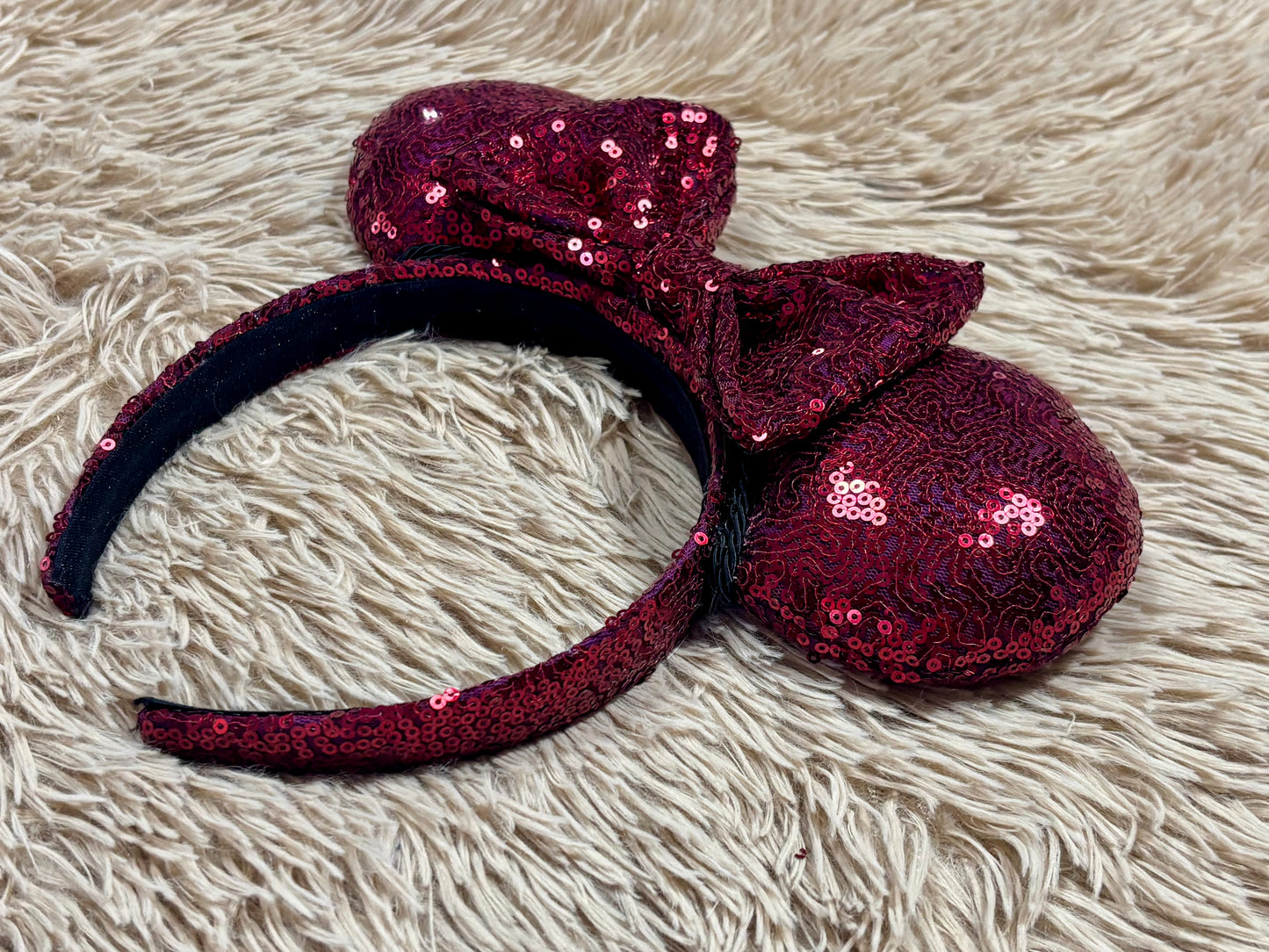 A sequined headband with large bow and ear-like shapes, covered in deep burgundy sequins and photographed on a cream shag rug.