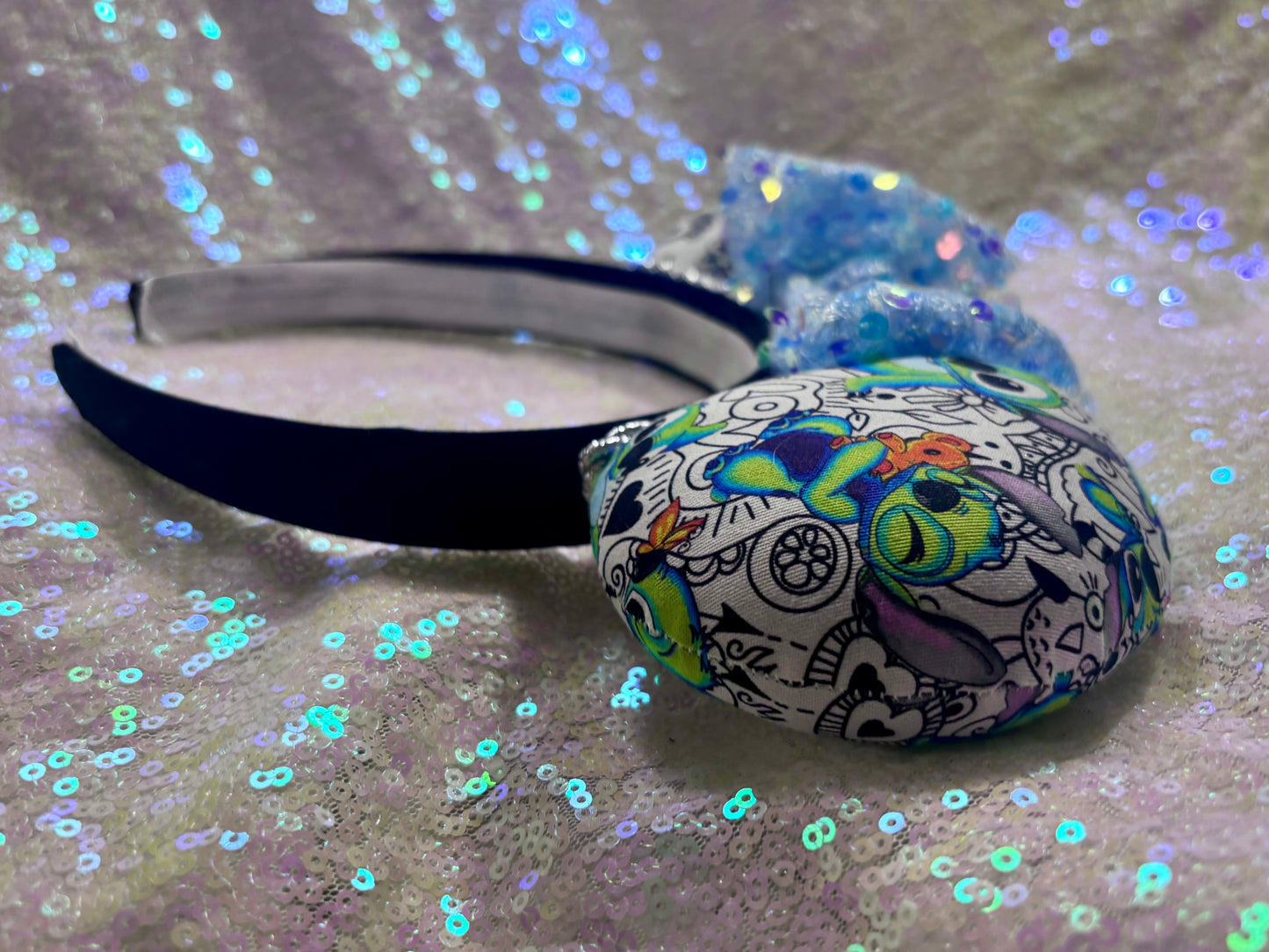 Side view of headband. Handmade and ready to ship Mouse ears with Stitch cartoon character design on the fabric with silver twisted trim on a black universal sized headband and a blue sequin bow.