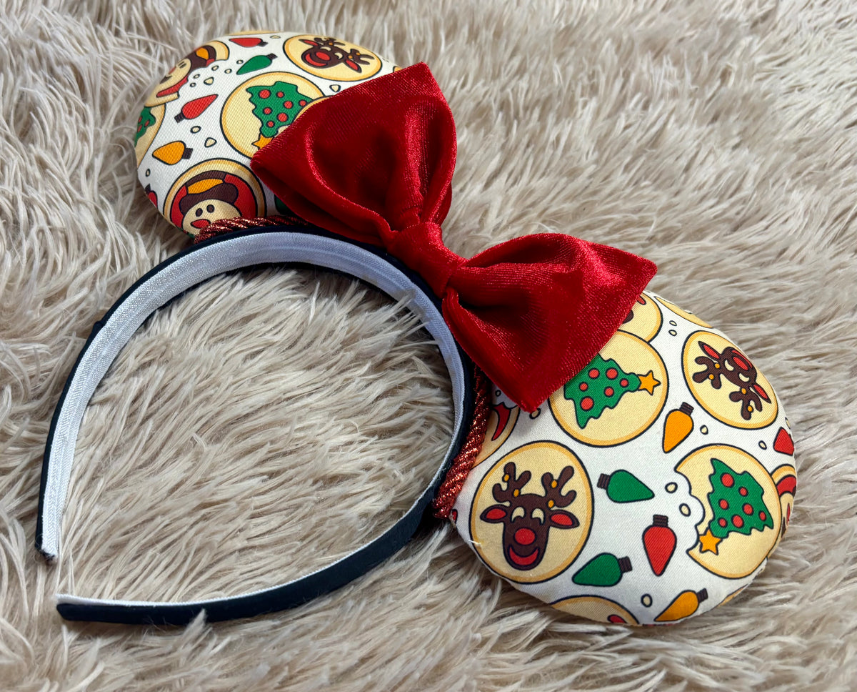 An ear headband featuring a festive Christmas print with red velvet bows, displayed on a beige faux fur background.