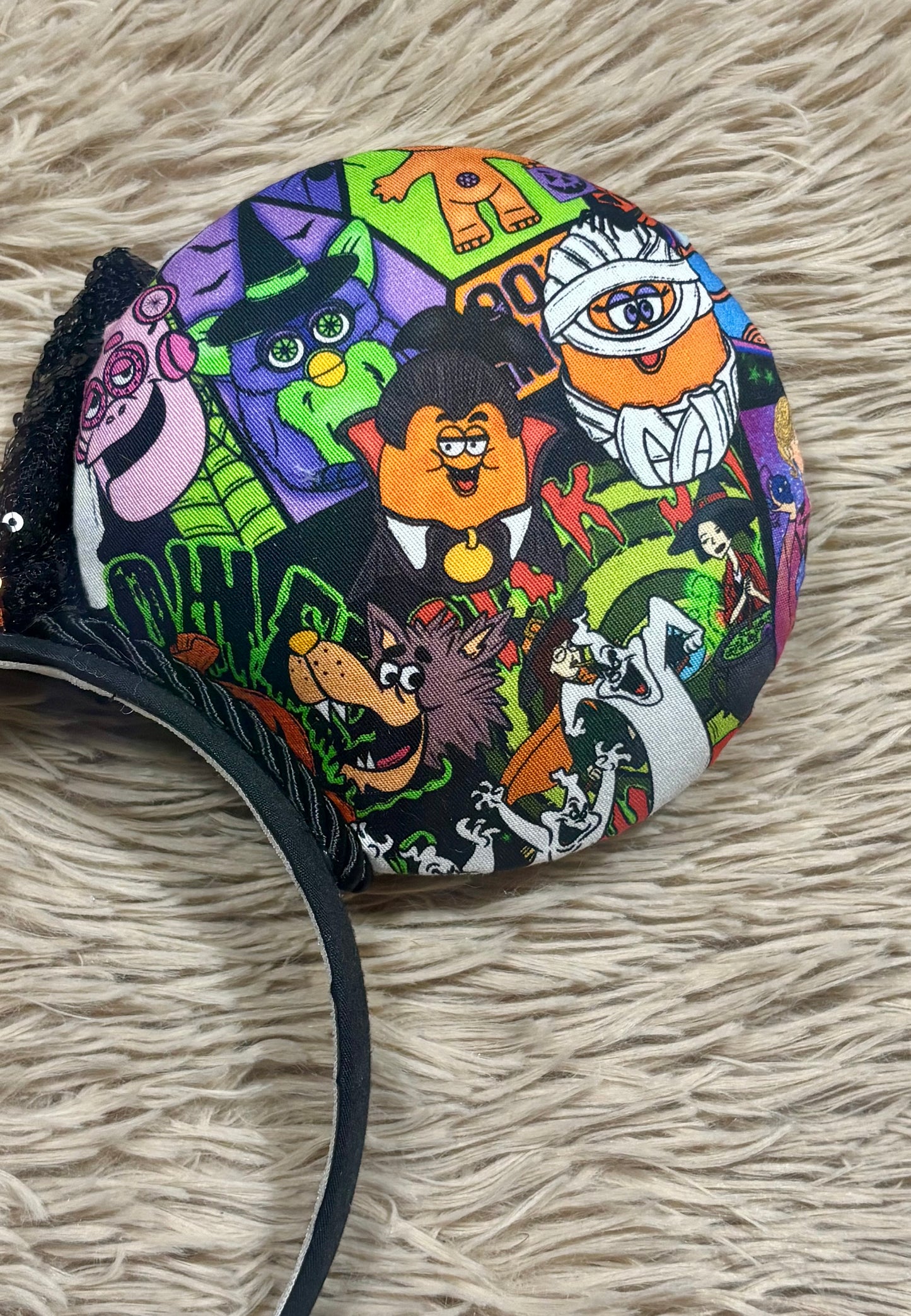 A round fabric-covered headband featuring a colorful Halloween-themed cartoon print with characters like a witch, vampire, mummy, and ghost on a black background, displayed on a beige faux fur surface.