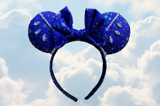 Blue glittery mouse ears headband with star accents and rhinestones on the ears with rhinestone trim. Shown on a cloud background.