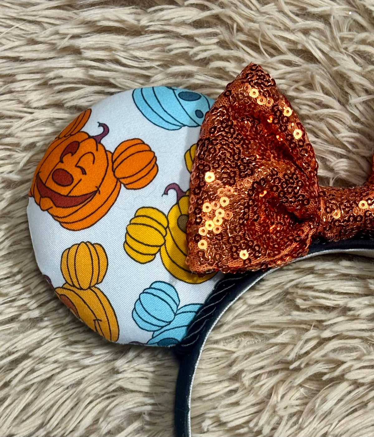 A close-up of an ear headband featuring a light blue fabric face with orange and yellow pumpkins and blue pumpkins, topped with a large orange sequined bow, resting on a beige shag rug.