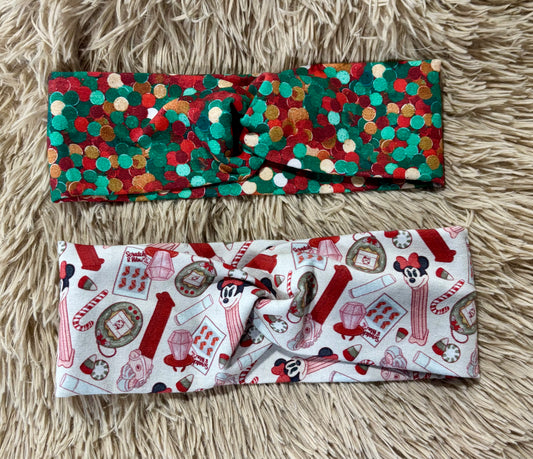 Two fabric headbands displayed flat on a beige faux-fur surface. The top headband has a multicolor holiday pattern with red, green, gold, and white polka dots. Bottom headband is nostalgic stocking stuffers.