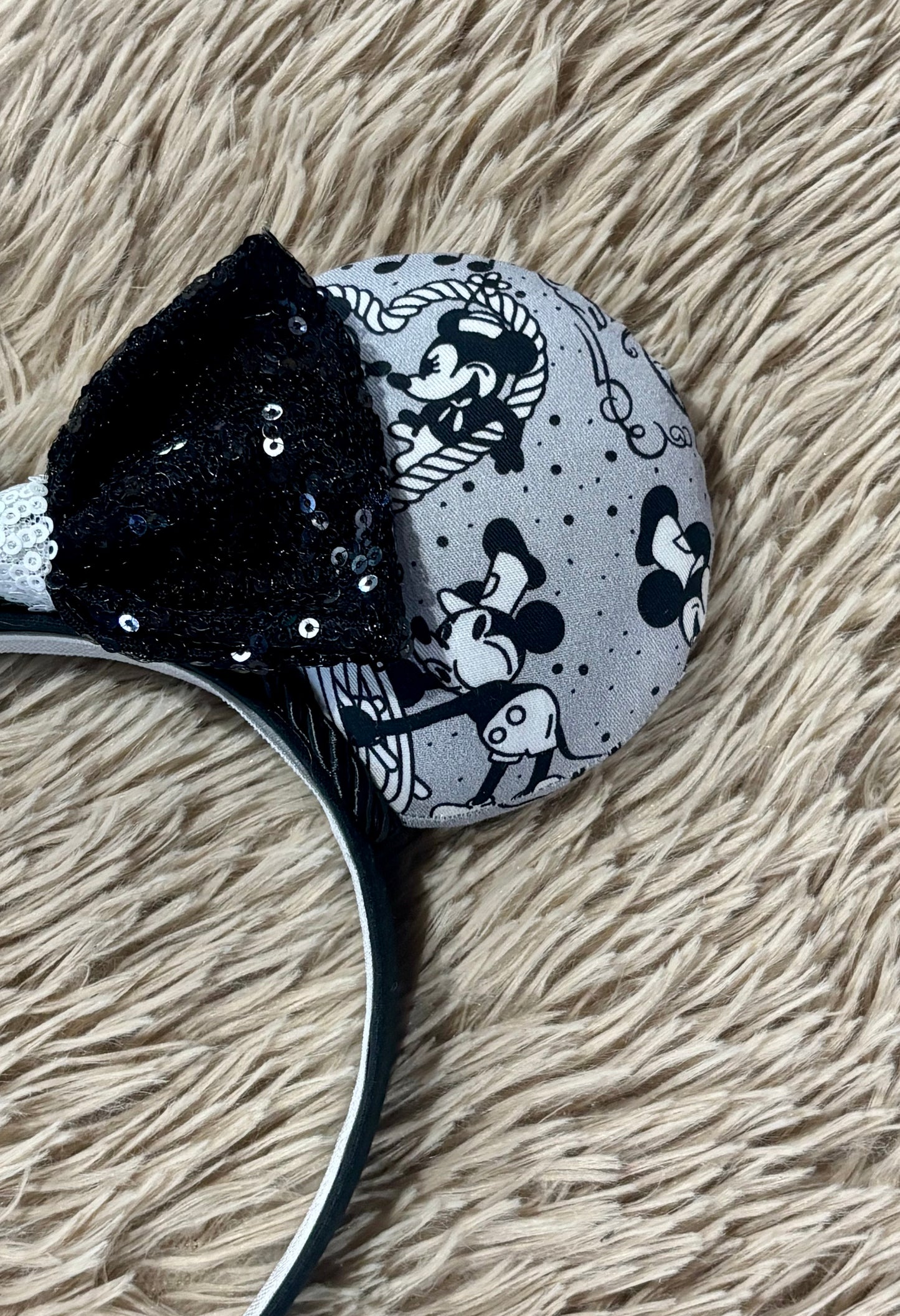 A pair of Mouse-themed ear headbands featuring a black and white patterned fabric with sequins, a black sequined bow with a white sequin middle, and a white headband, displayed on a beige faux fur background.