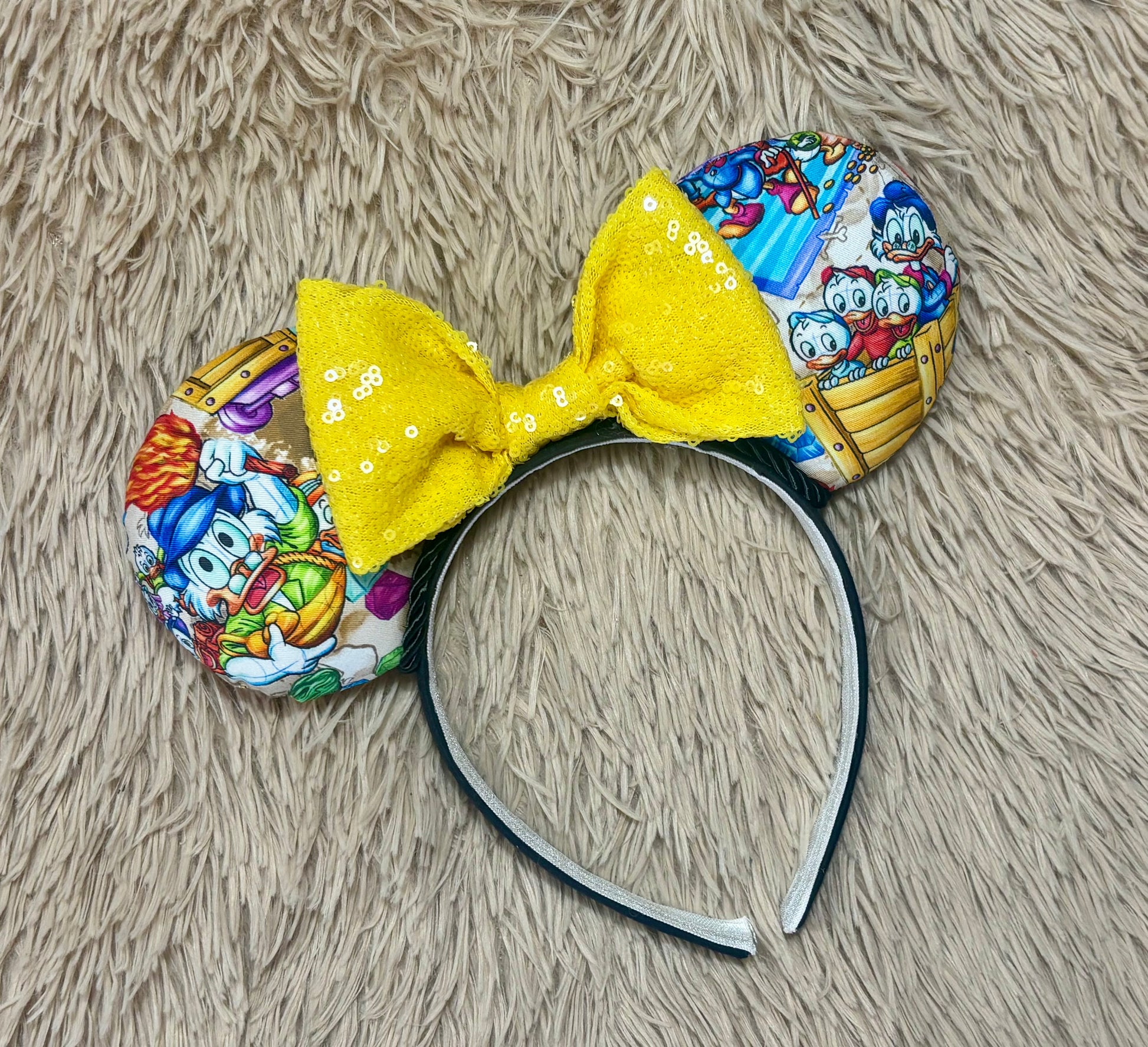 A headband featuring a colorful cartoon print of famous cartoon Duck characters, accented by a large yellow sequined bow in the center.