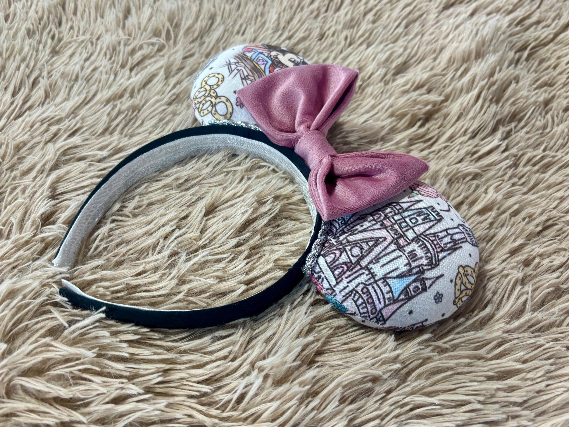 A pair of handmade ear headbands with a large dusty pink velvet bow, featuring printed park themed patterns on the ears and resting on a beige faux fur surface.