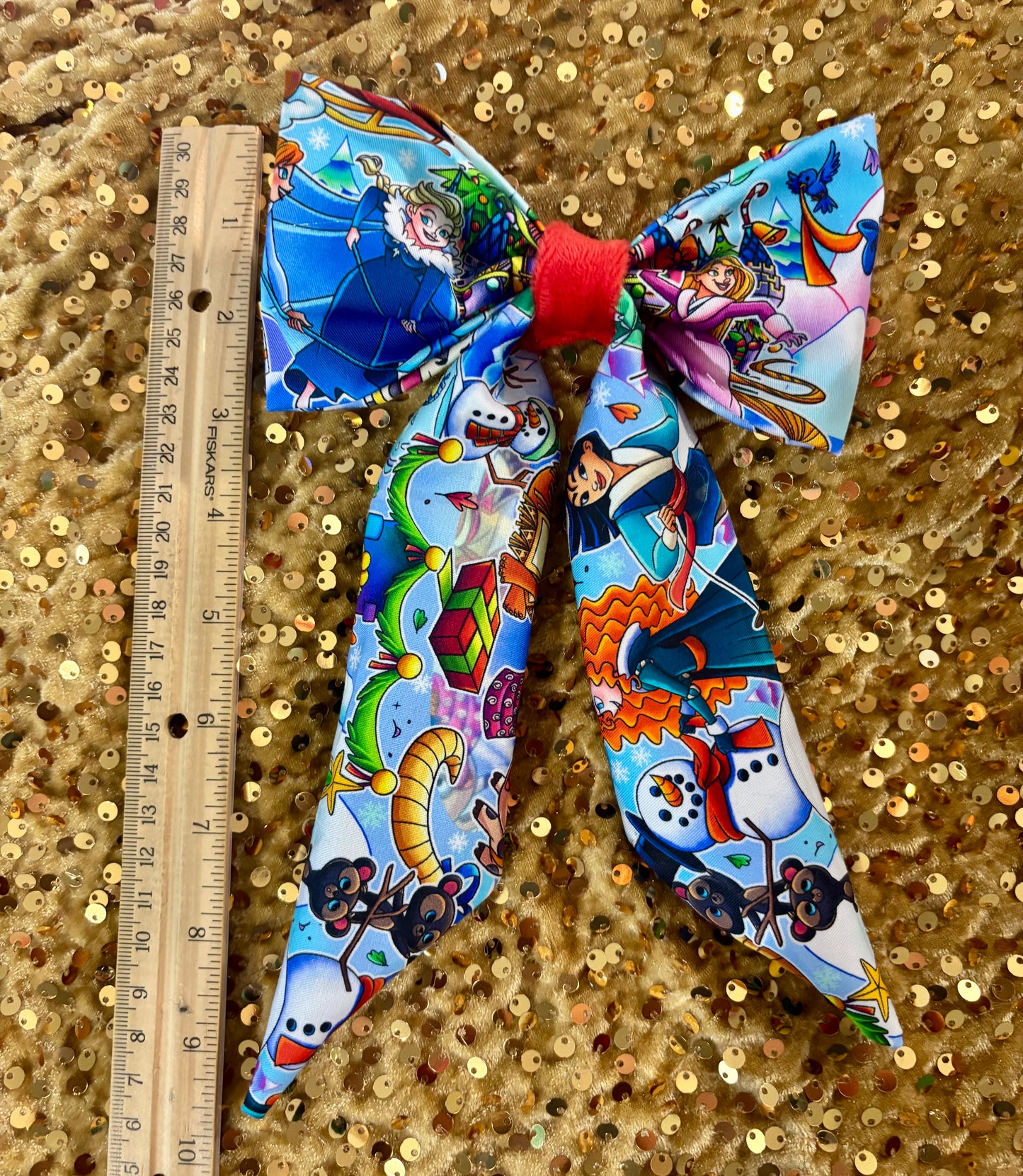 Colorful bow with Princess characters on a gold sequin background, next to a ruler for scale.