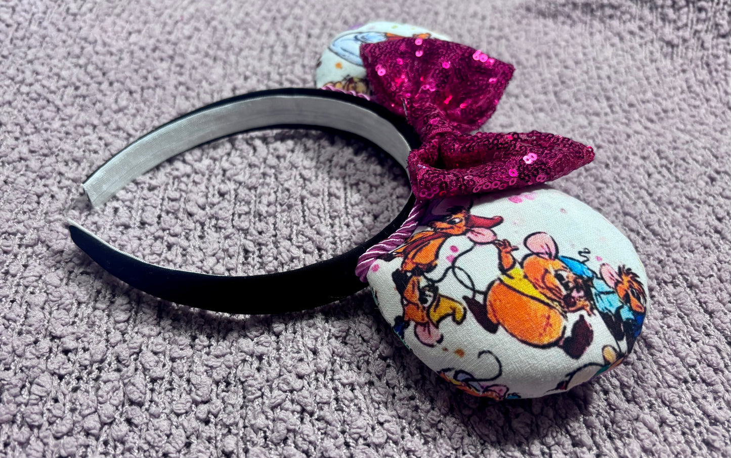 Side view of mouse ears Handmade and ready to ship Sewing Mice mouse ears with the fabric on both the front and back. The headband is black universal sized and fits most. The set has a hot pink sequin bow with pink twisted trim along the inner mouse ears. Item is shown on a lavender fluffy background.
