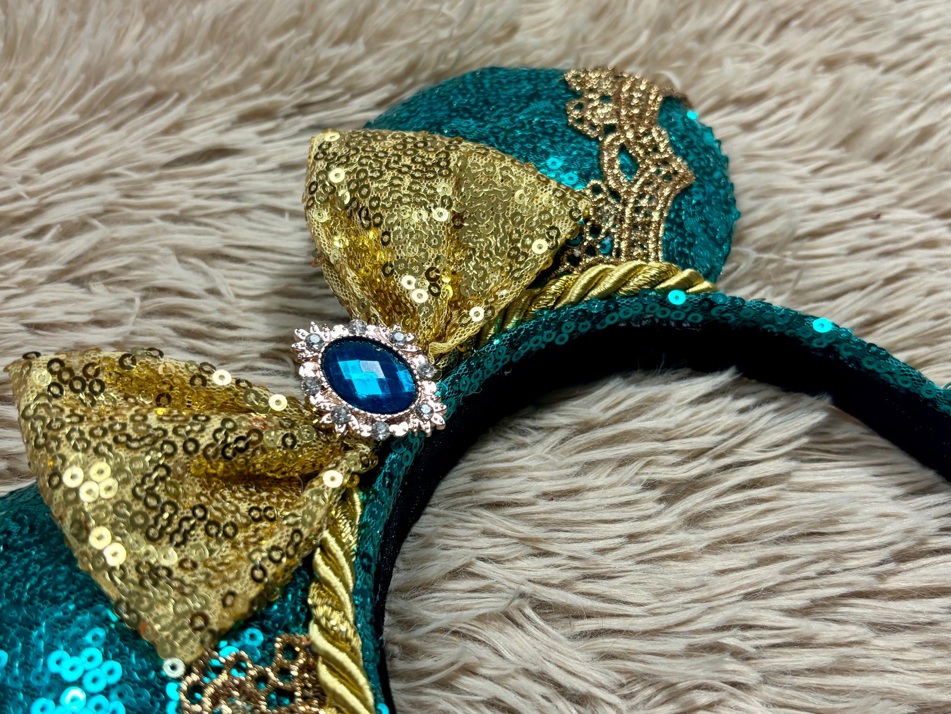A close-up of a teal and gold sequined headband with a large gold sequin bow and a central blue rhinestone, resting on a gray faux fur surface.
