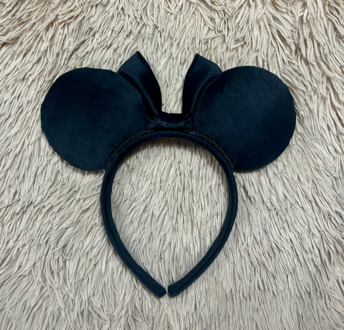 Black velvet mouse ears headband with a bow, displayed on a textured beige faux fur background. Shown from the back side view of set.