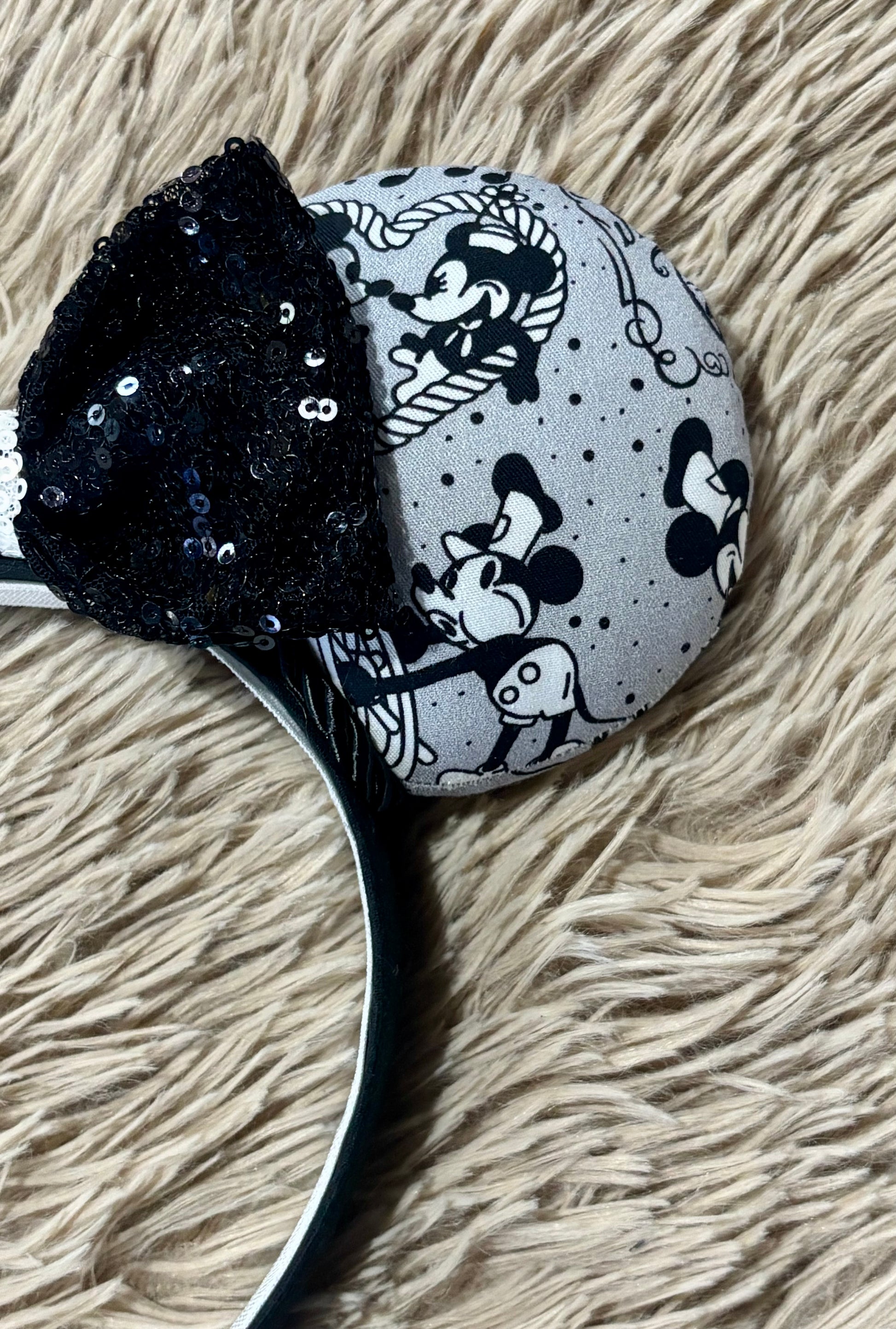 A pair of Mouse-themed ear headbands featuring a black and white patterned fabric with sequins, a black sequined bow with a white sequin middle, and a white headband, displayed on a beige faux fur background.