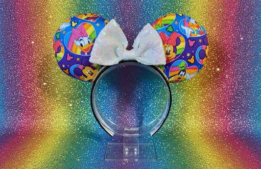 A pair of the ear headband covers with colorful cartoon print and a white bow, displayed on a clear stand against a rainbow glitter background.