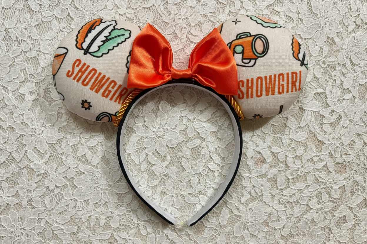 Showgirl-themed mouse ear headband with orange satin bow and twisted trim. Shown on a lace background