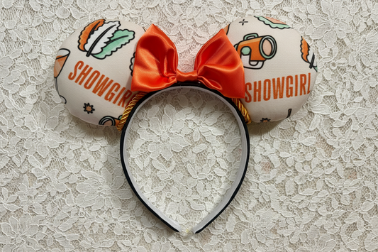 Showgirl-themed mouse ear headband with orange satin bow and twisted trim. Shown on a lace background