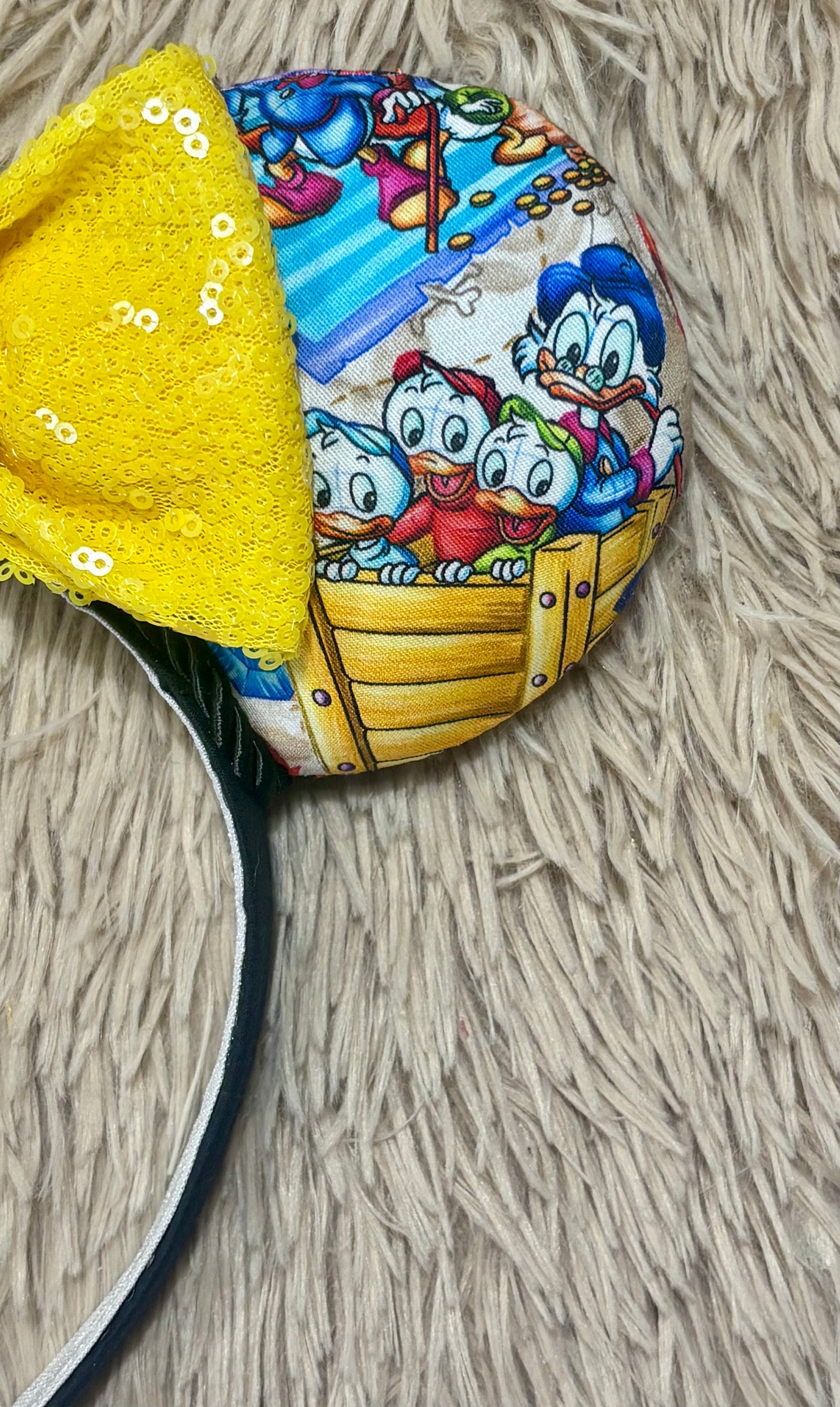 A headband featuring a colorful cartoon print of famous cartoon Duck characters, accented by a large yellow sequined bow in the center.