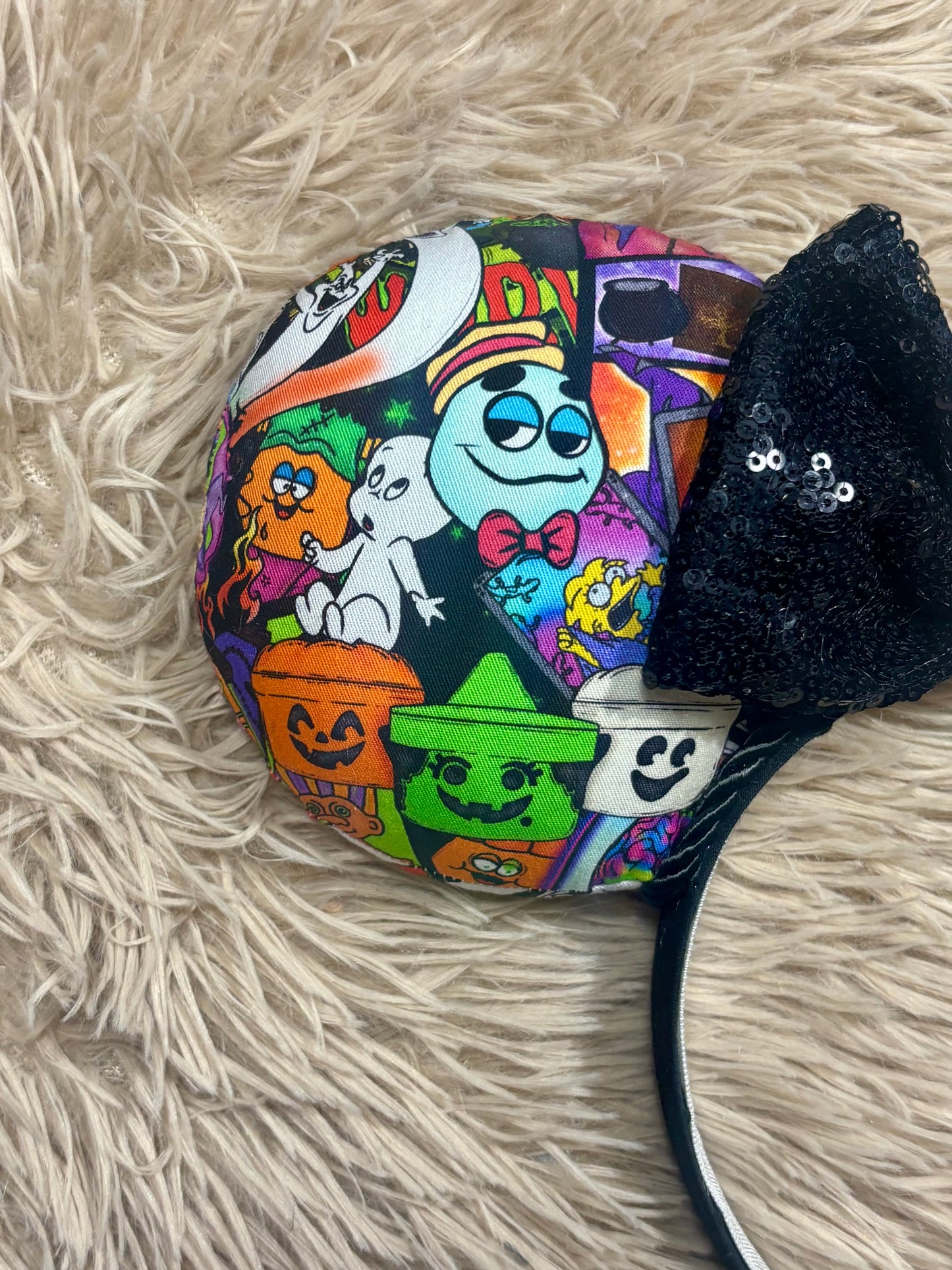 A mouse-shaped headband featuring a colorful Halloween-themed collage print with cartoon ghosts, pumpkins, and a witch hat, finished with a black sequined cat-ear accent on a beige faux fur background.