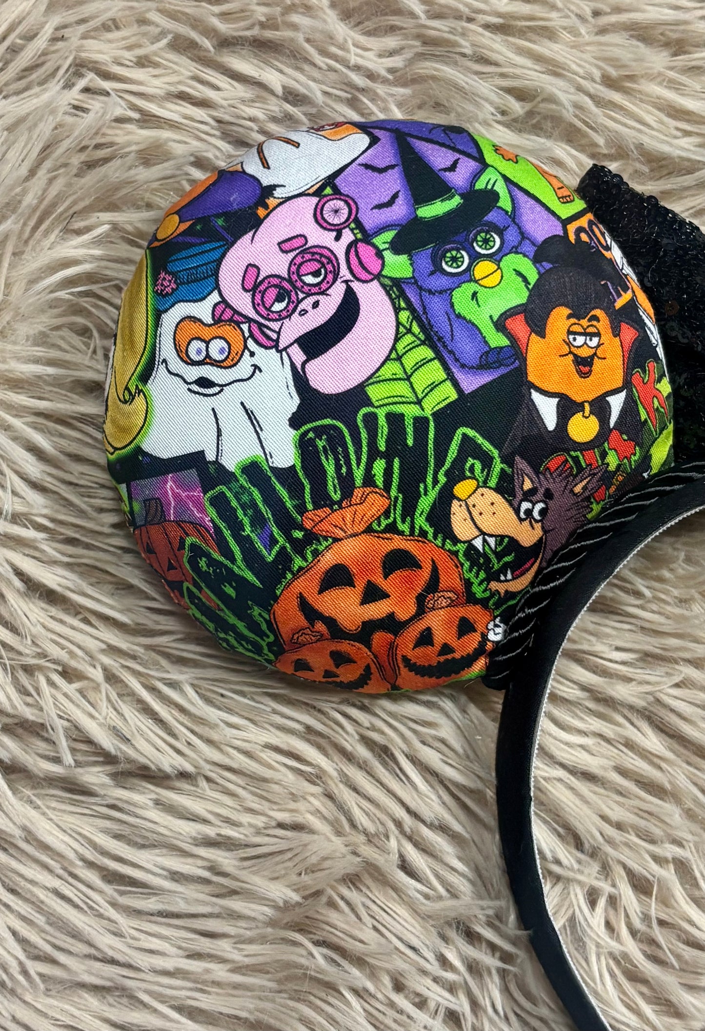 An ear headband accessory featuring a colorful Halloween-themed print on the outer ears and a black glitter bow in the center with black twisted trim along the inner ears. Close up of backside of ear on headband.