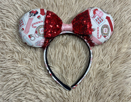 Christmas themed ears headband featuring a festive holiday print with red and white candy cane motifs, a red sequined bow, and a black headband, displayed on a beige faux fur background.