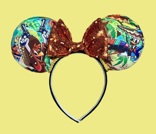 A pair of cabellero inspired ears headband featuring colorful cartoon character prints on the ears and a large sequined copper-brown bow on top, shown on a yellow background.